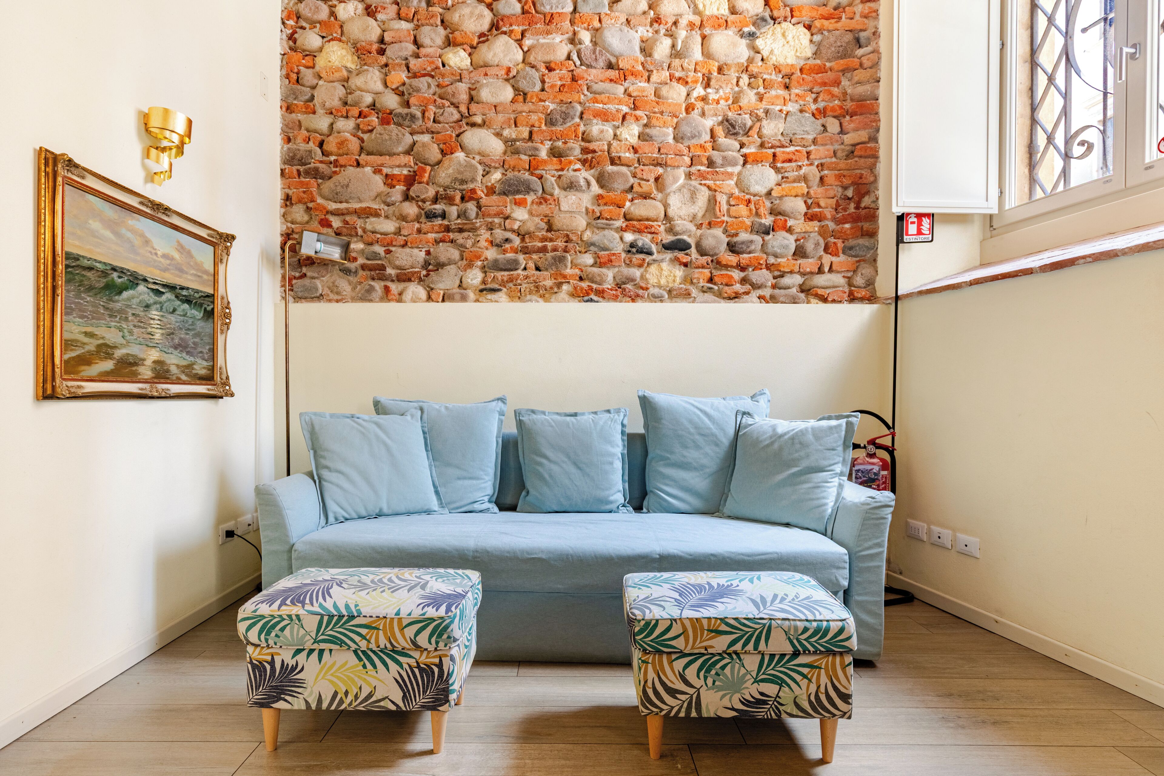 A living room with a blue sofa, two patterned ottomans, a stone wall, a framed painting, and a wall-mounted light fixture.