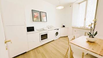 Une cuisine compacte avec des meubles blancs, un four à micro-ondes, un four et un lave-linge. On y trouve une table à manger avec des chaises et une plante en pot sur le rebord de la fenêtre.