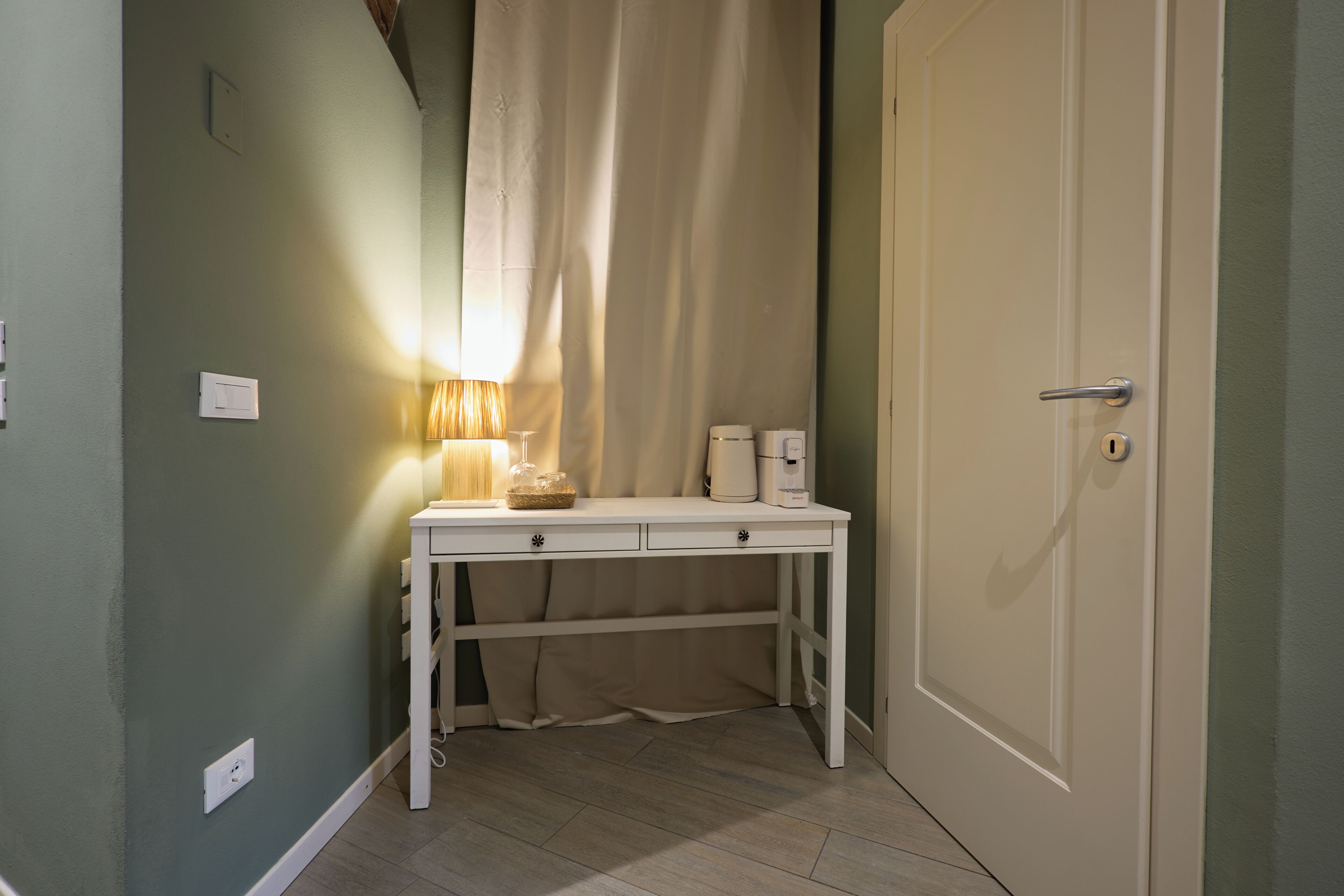 A small room with a desk, a lamp, a coffee maker, and a door.
