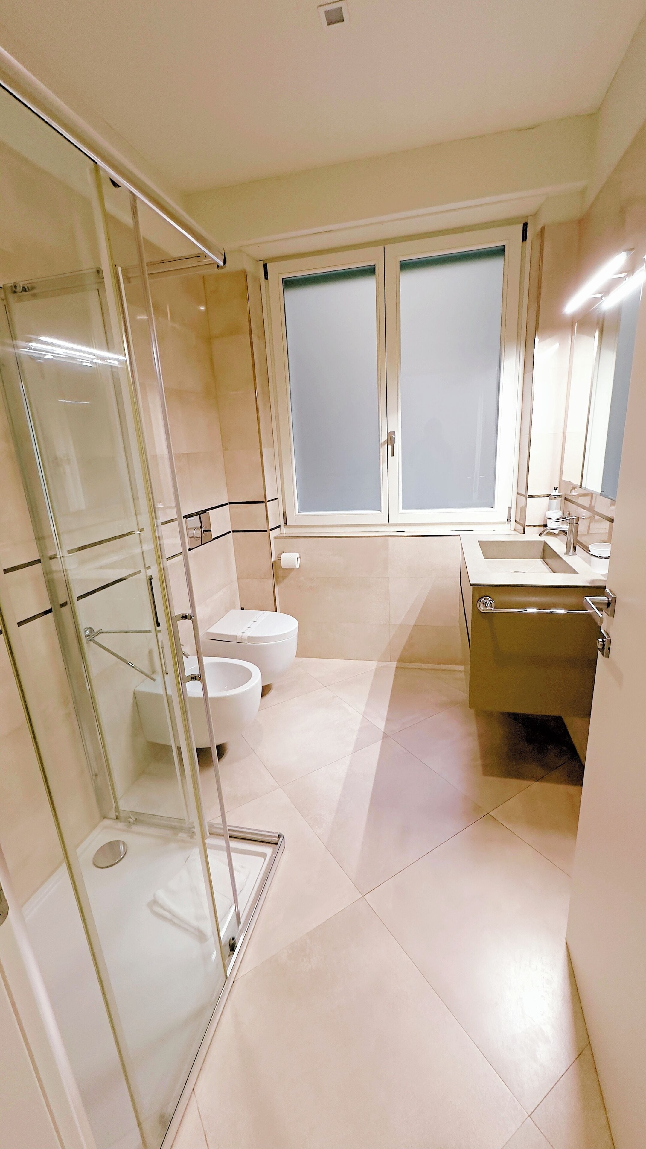 A modern bathroom with a glass shower enclosure, a toilet, a sink, and a window with frosted glass.