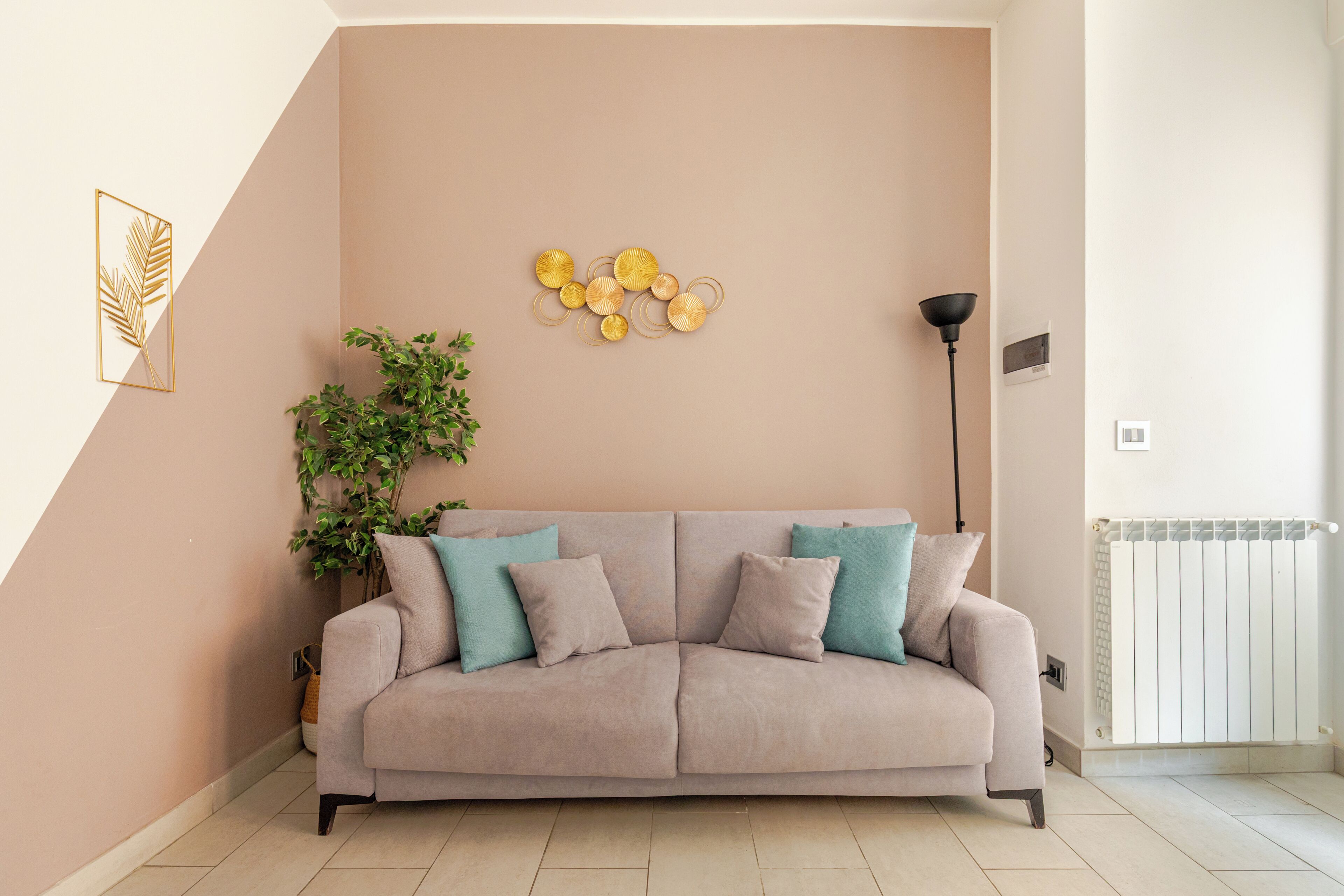 Un sofá color beige con cojines verde azulado, una planta en maceta y cuadros en una habitación con radiador.