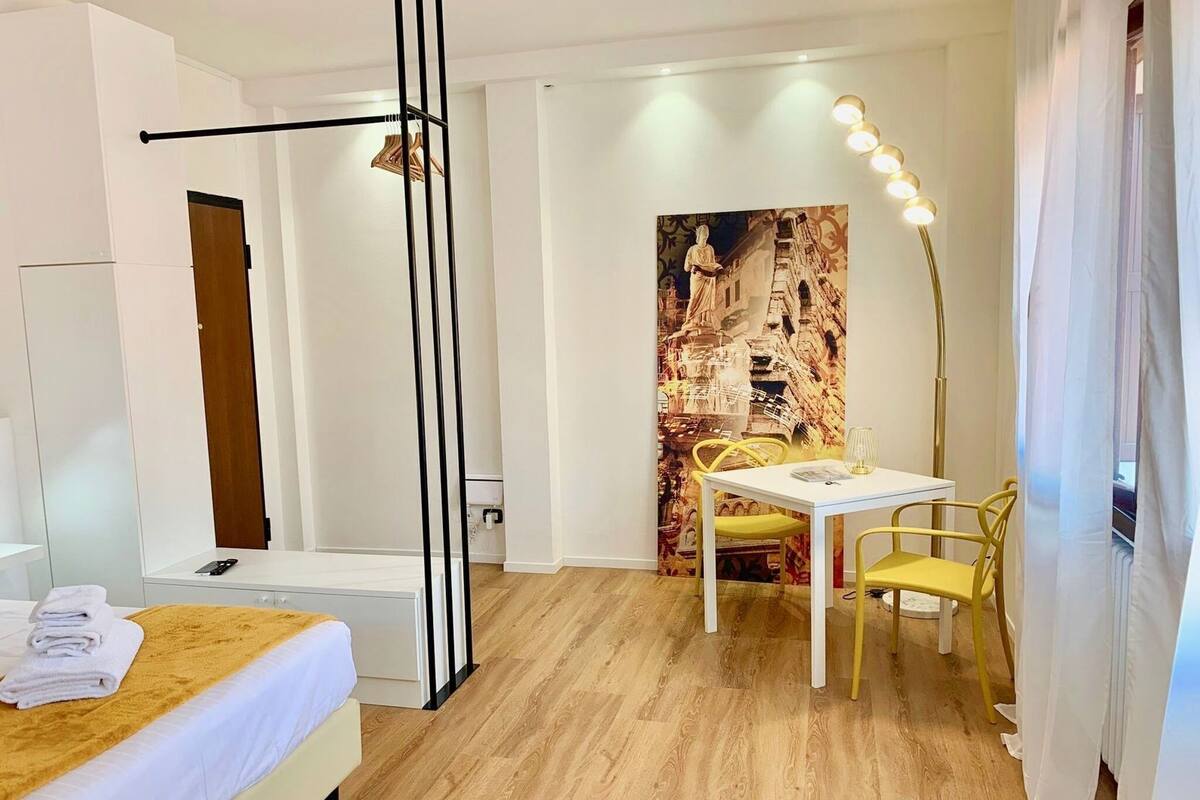 A hotel room with a bed, a desk with a chair, a standing lamp, and a wall with a large painting.