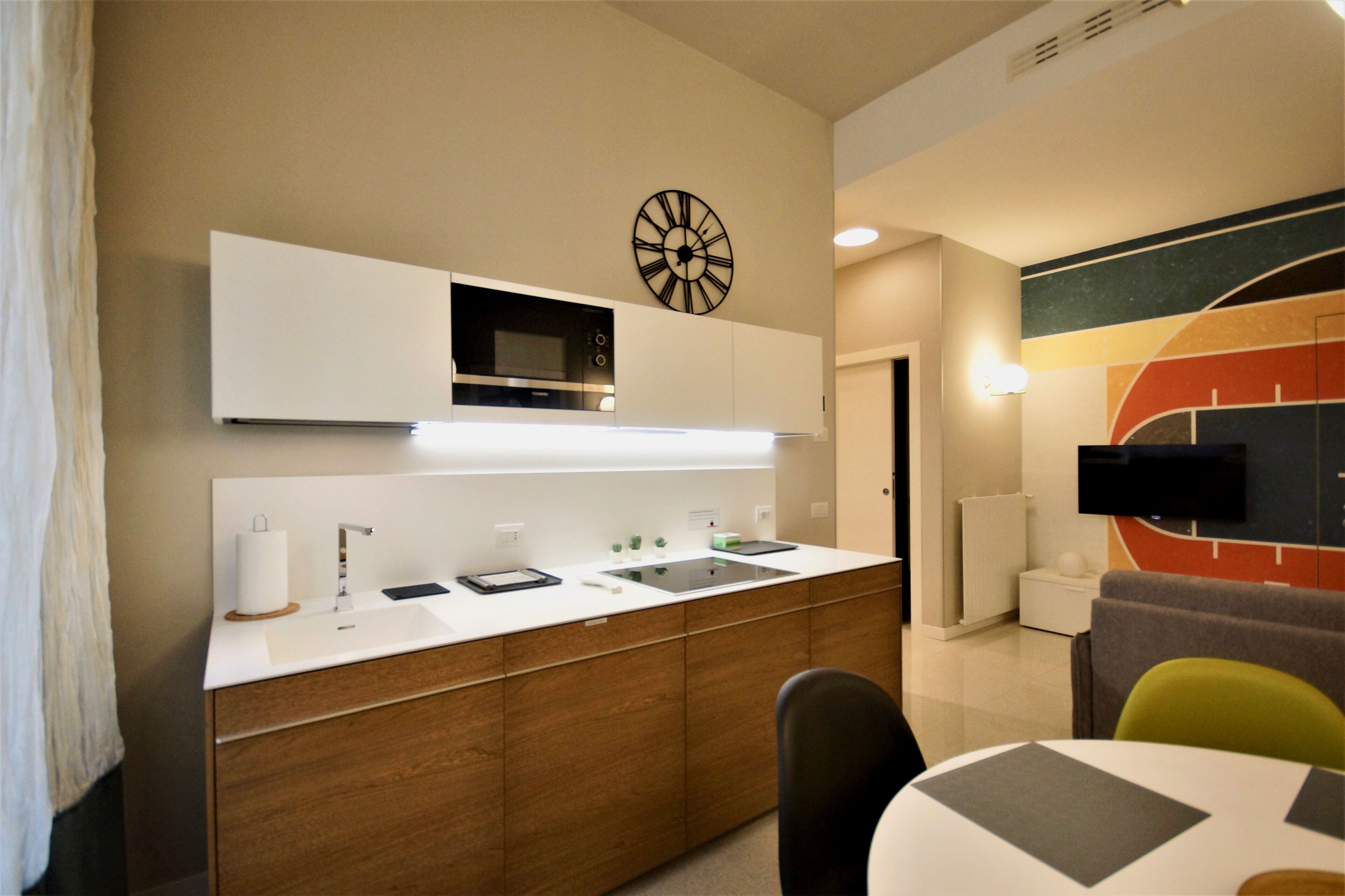 A modern kitchen with a microwave, sink, and wooden cabinets. There is a clock on the wall and a TV mounted above the counter.
