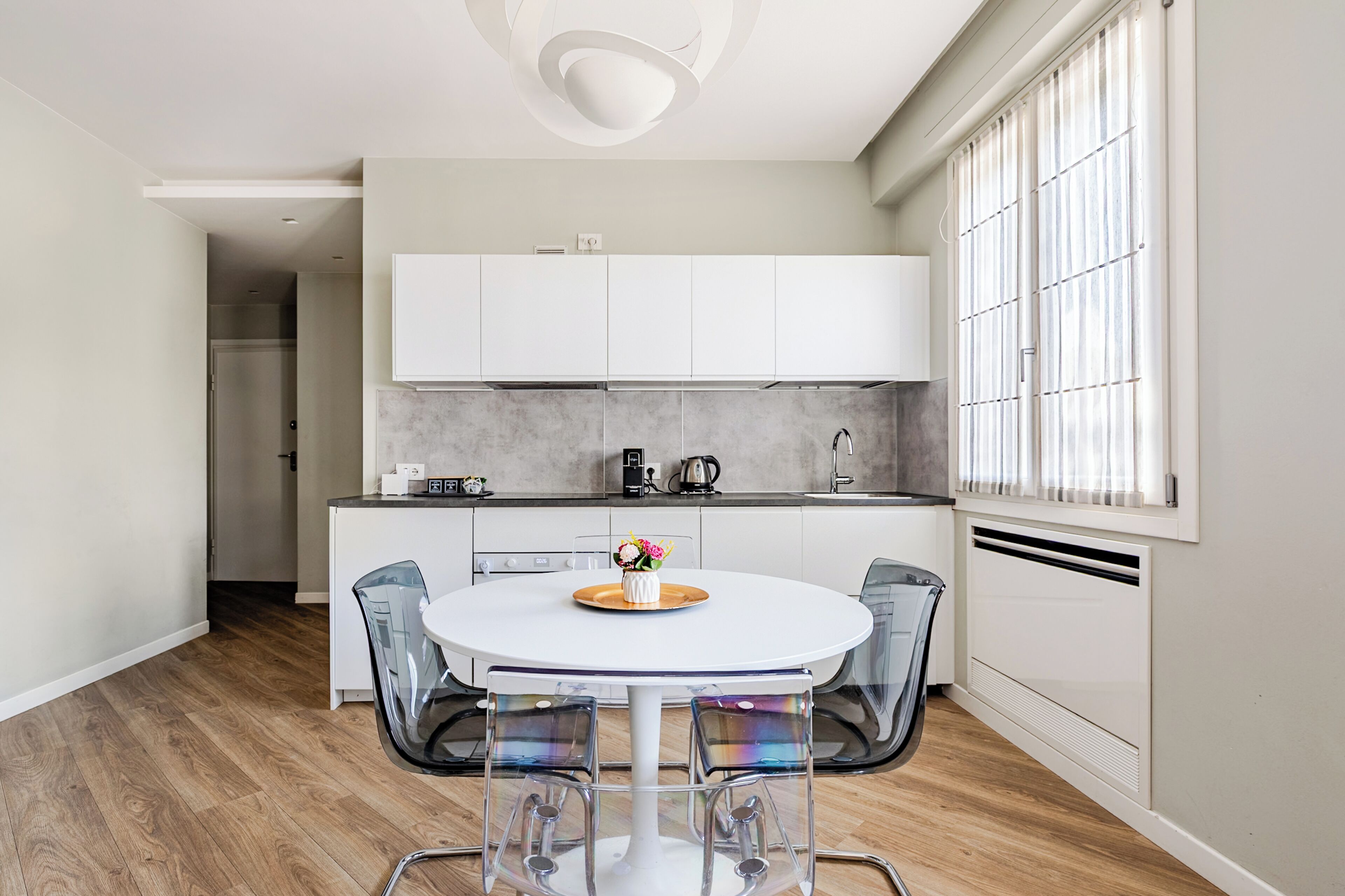 Una cocina moderna con armarios blancos, una mesa redonda de comedor con sillas transparentes y una ventana con persianas.