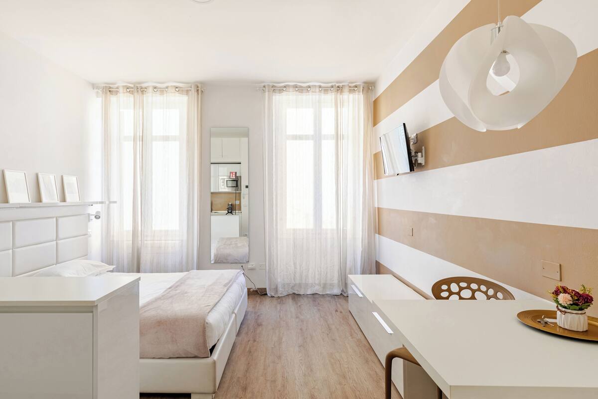 A modern hotel room with a bed, a desk, a chair, a dining table, and a kitchen area in the background.