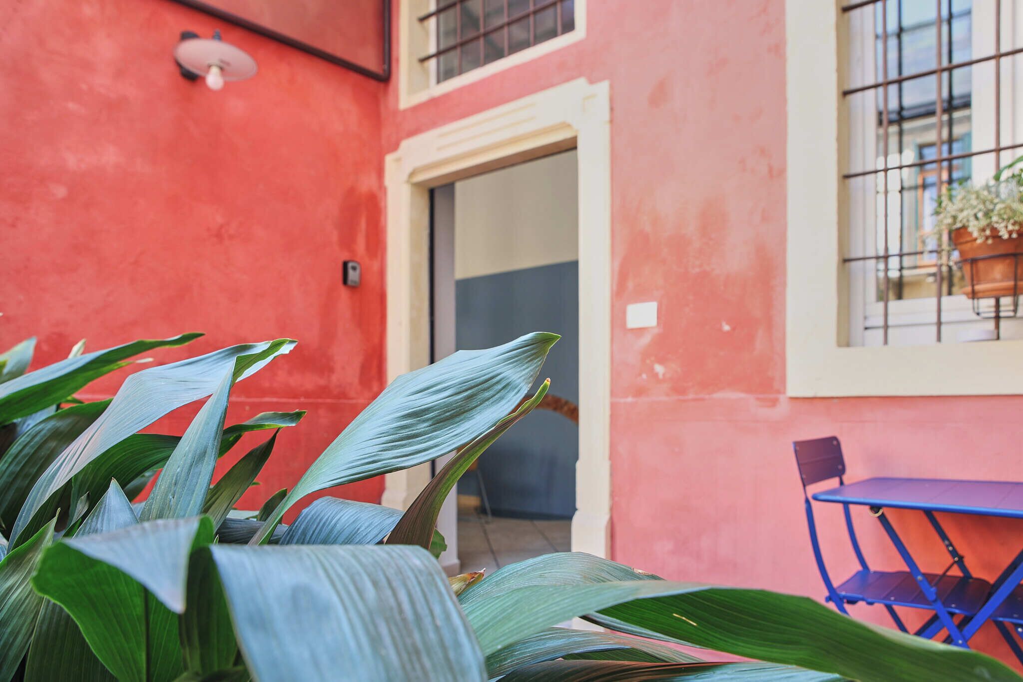 A red building with a door, a window with a plant, and a blue table and chair.