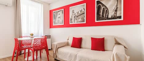 Ein Zimmer mit einem weißen Sofa, roten Sesseln, einem Esstisch und drei schwarz-weißen Fotografien an der Wand.