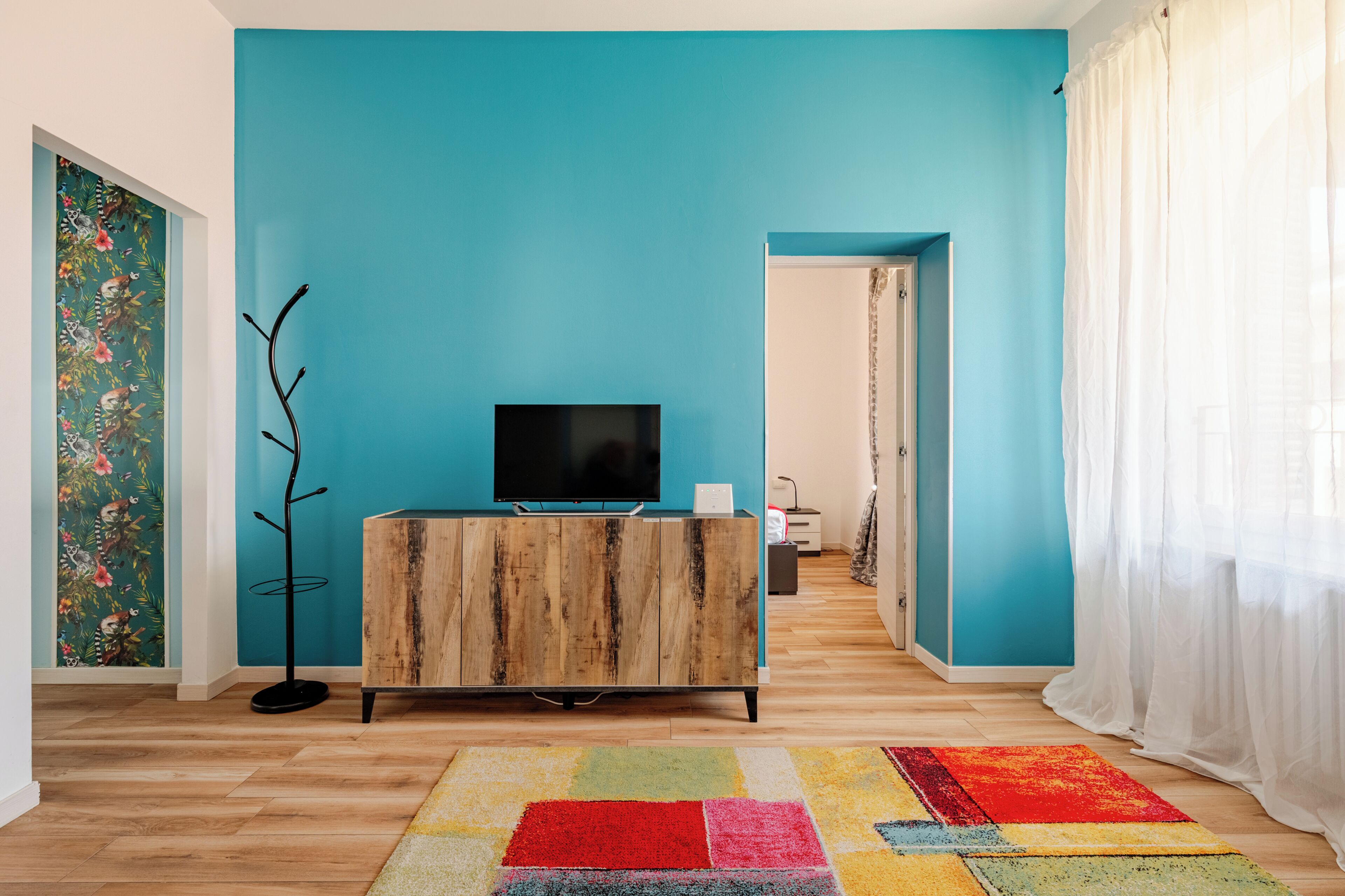 Una habitación con pared verde azulado, un mueble de madera, televisor de pantalla plana, alfombra colorida y una puerta que conduce a otra habitación.