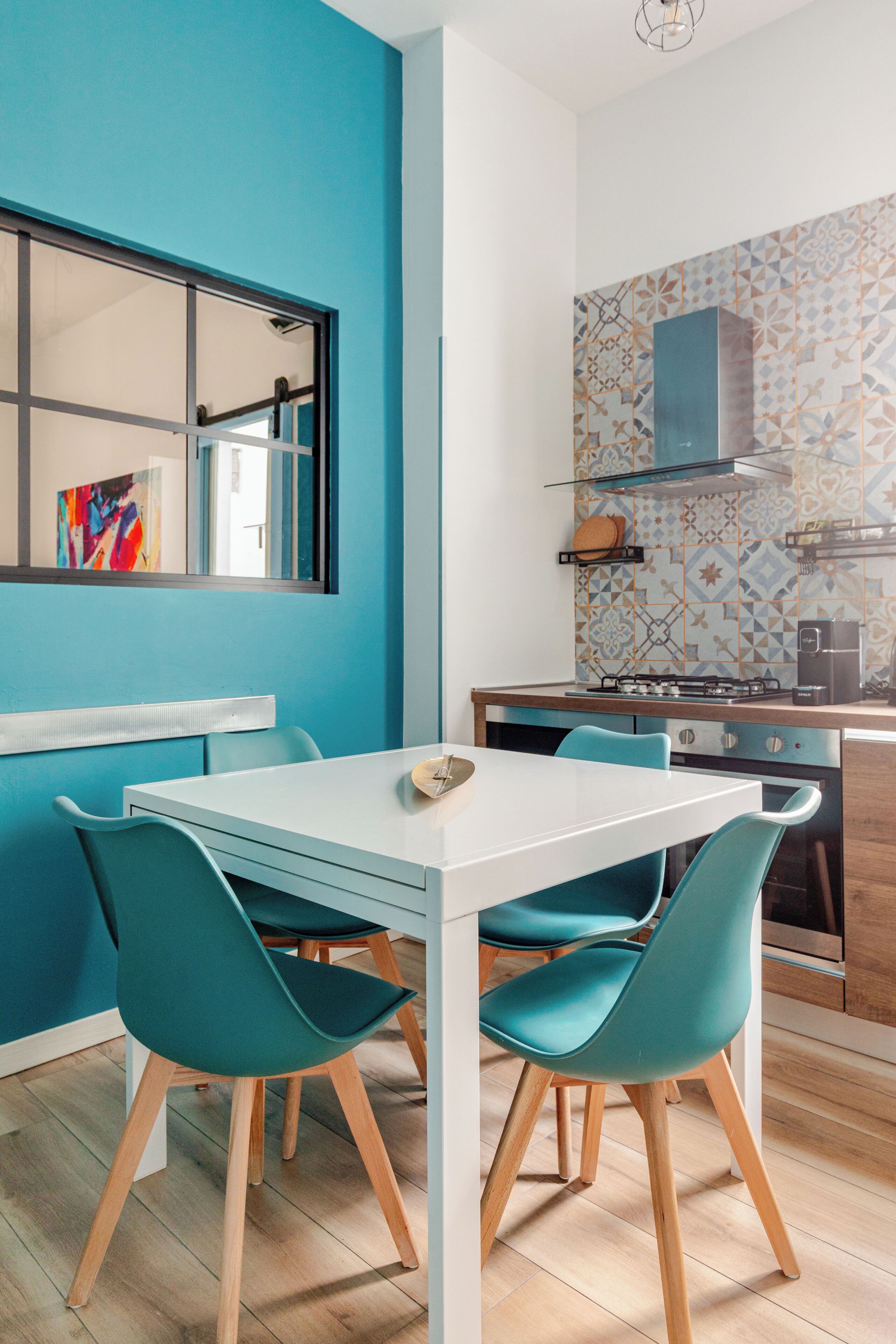 A modern kitchen with a white table and teal chairs, a window with a colorful painting, and a patterned backsplash.