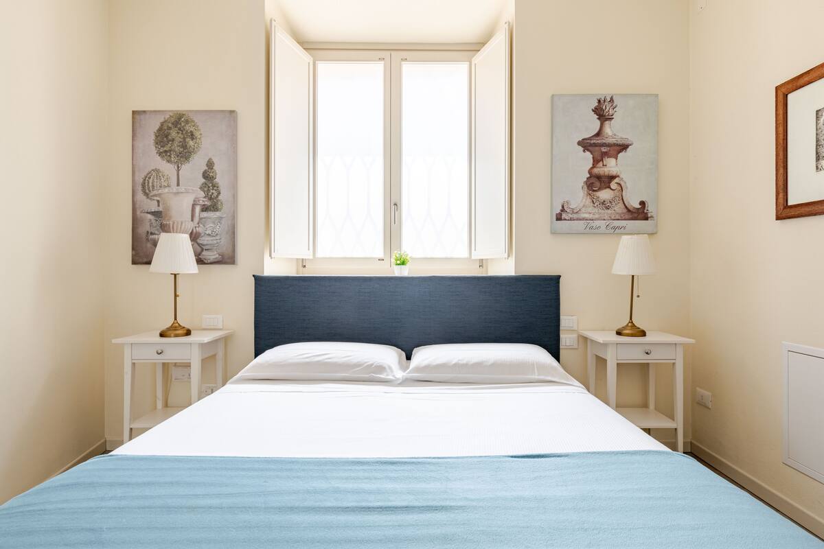 A bedroom with a bed, two bedside tables, a window, and two wall-mounted lamps.