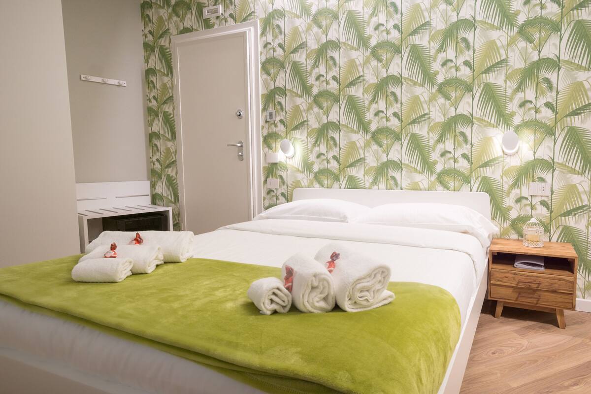 A hotel room with a bed, a wooden nightstand, a mirror, and a wall with a tropical leaf pattern.