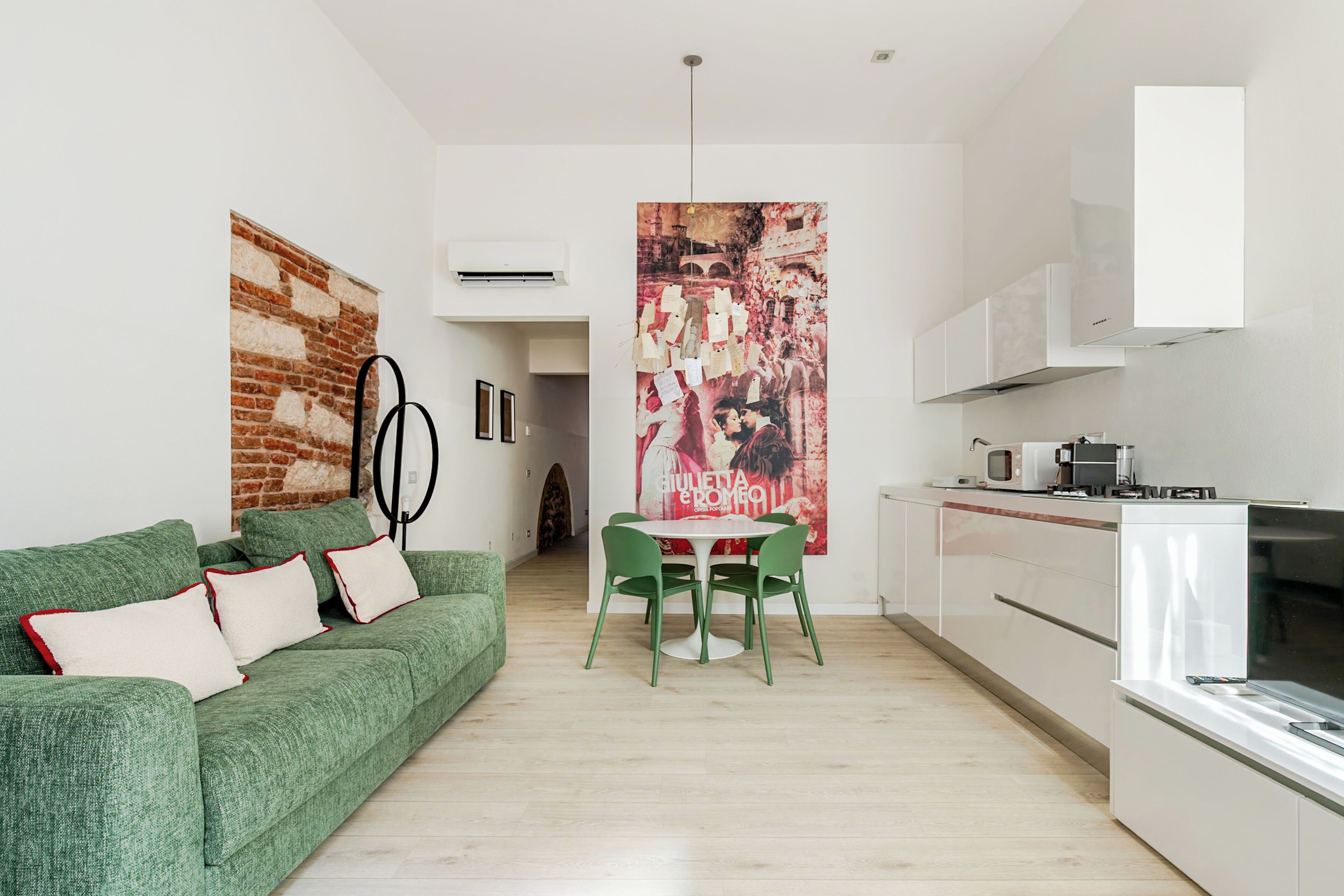 A modern living room with a green sofa, a dining table with chairs, a kitchen area with white cabinets, and a large poster on the wall.