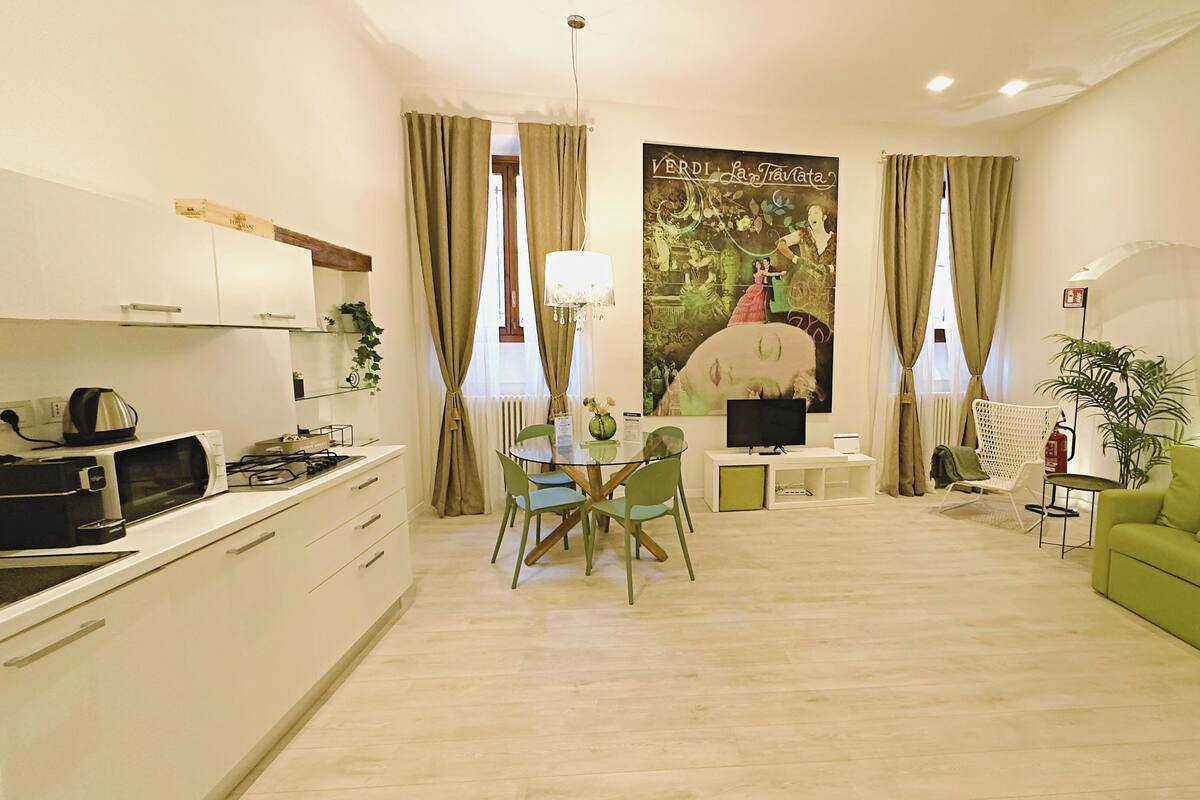A modern living room with a green sofa, a dining table with chairs, a kitchen area with a microwave, and a TV.