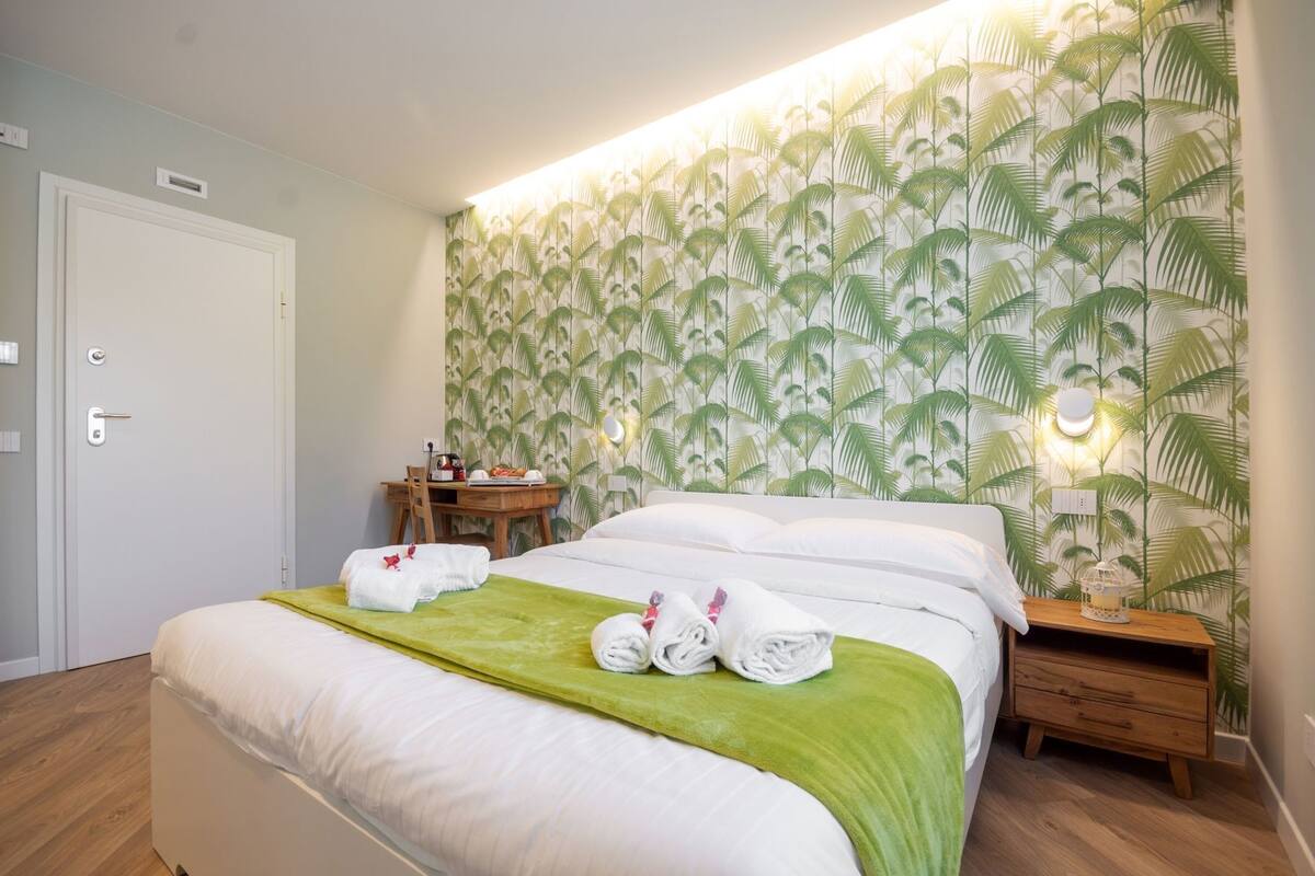 A hotel room with a bed, a wooden nightstand, a desk with a chair, and a wall with a leaf pattern.