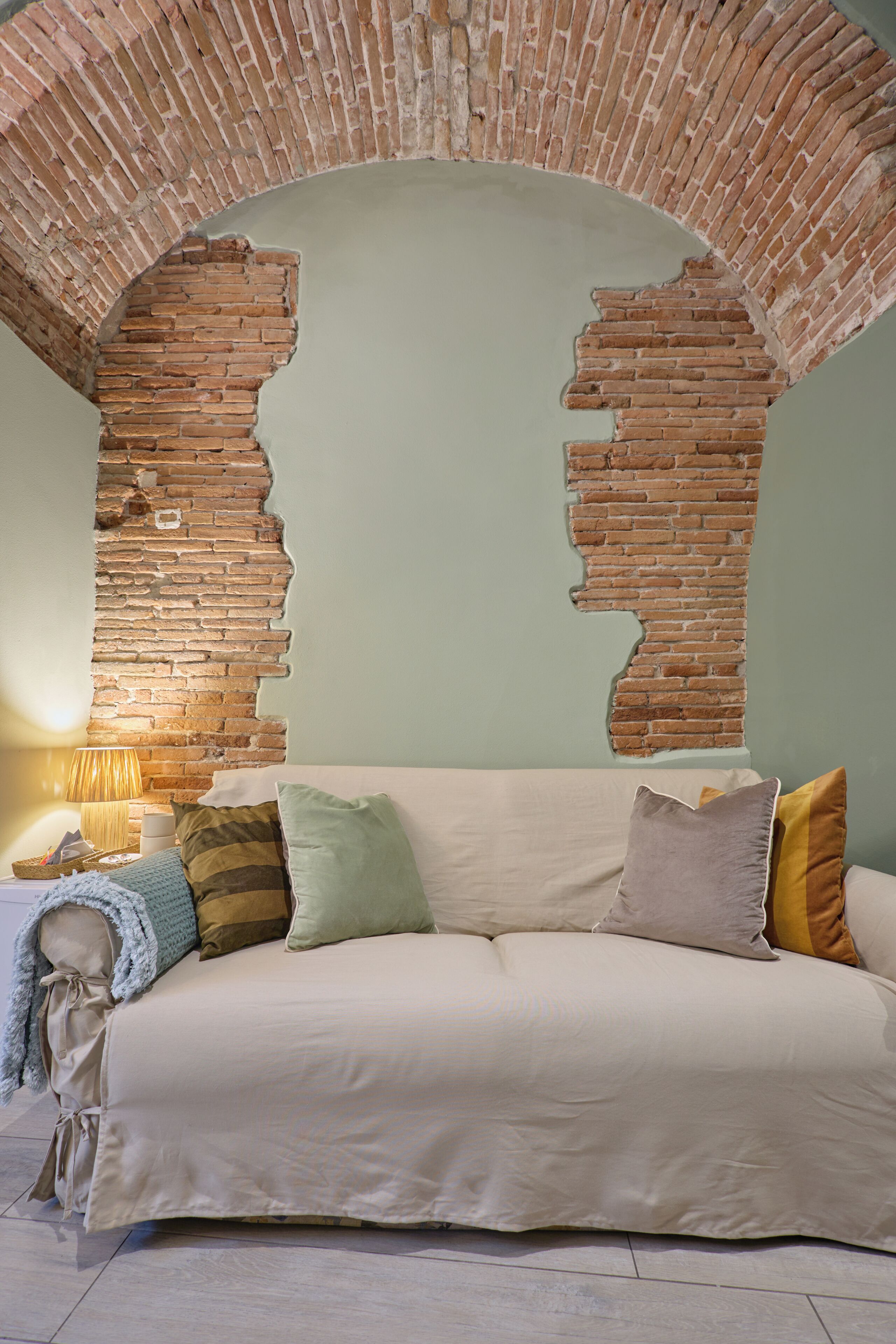A room with a brick archway, a sofa with cushions, a lamp, and a blanket.