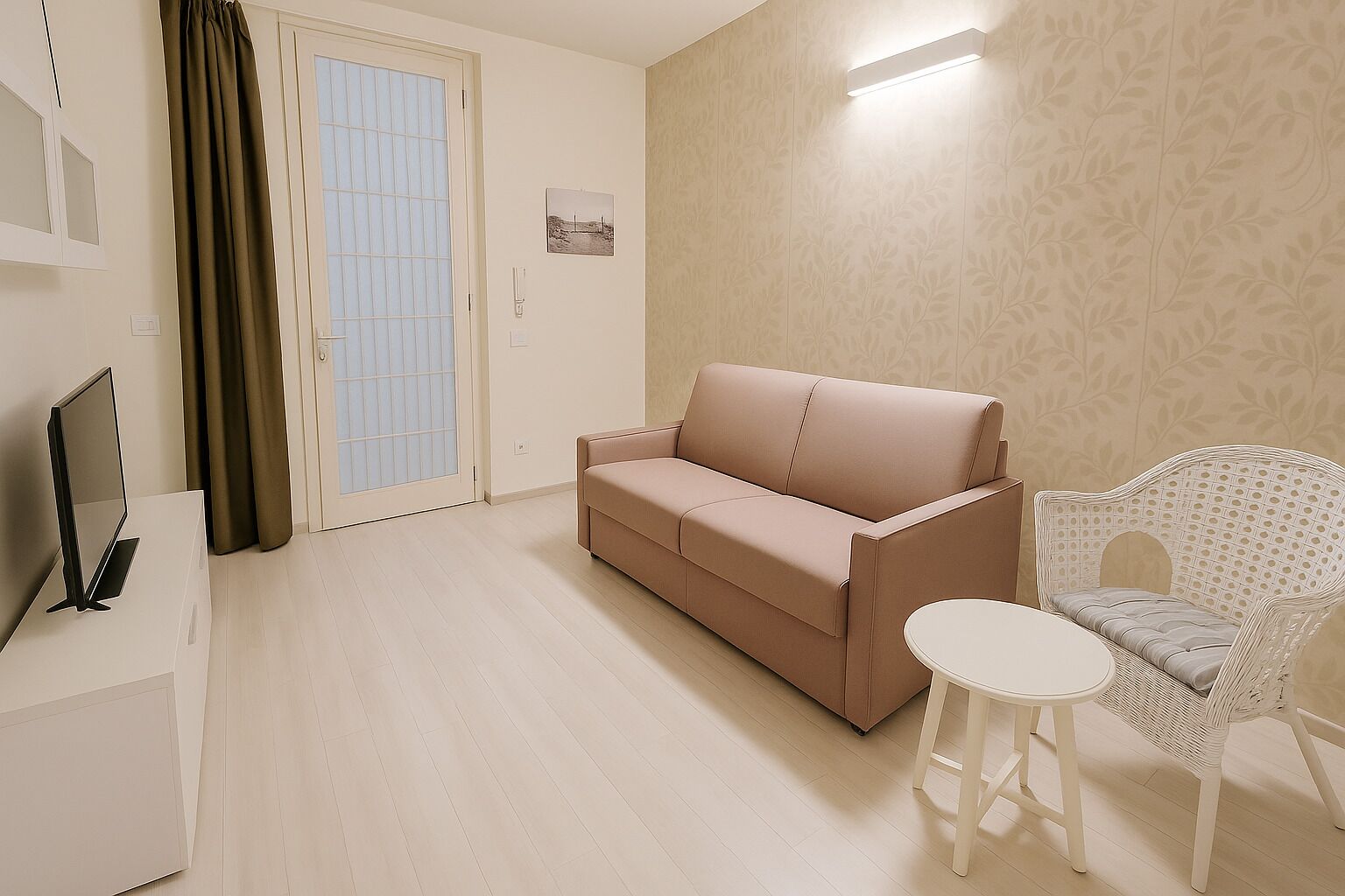 A living room with a beige sofa, a wicker chair, a small white table, a TV on a stand, and a door with a frosted glass panel.