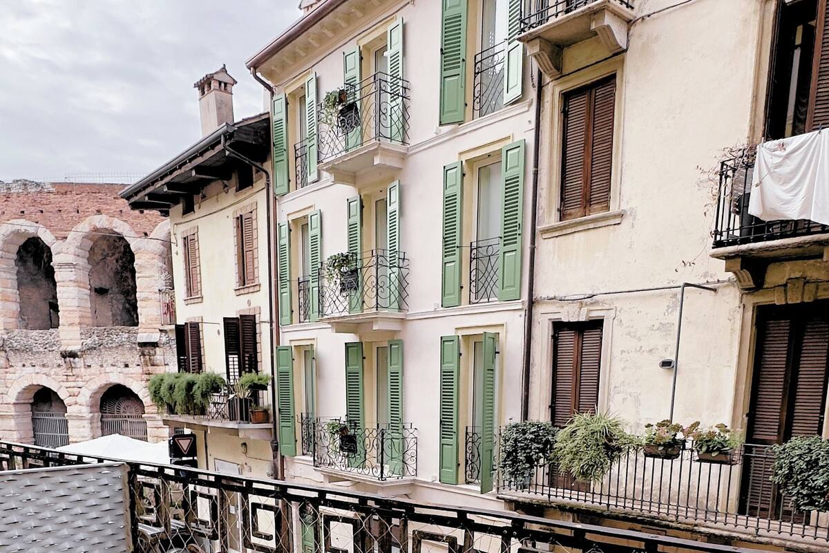 An outdoor patio with a round table and chairs, a metal fence, and buildings with green shutters and balconies in the background.