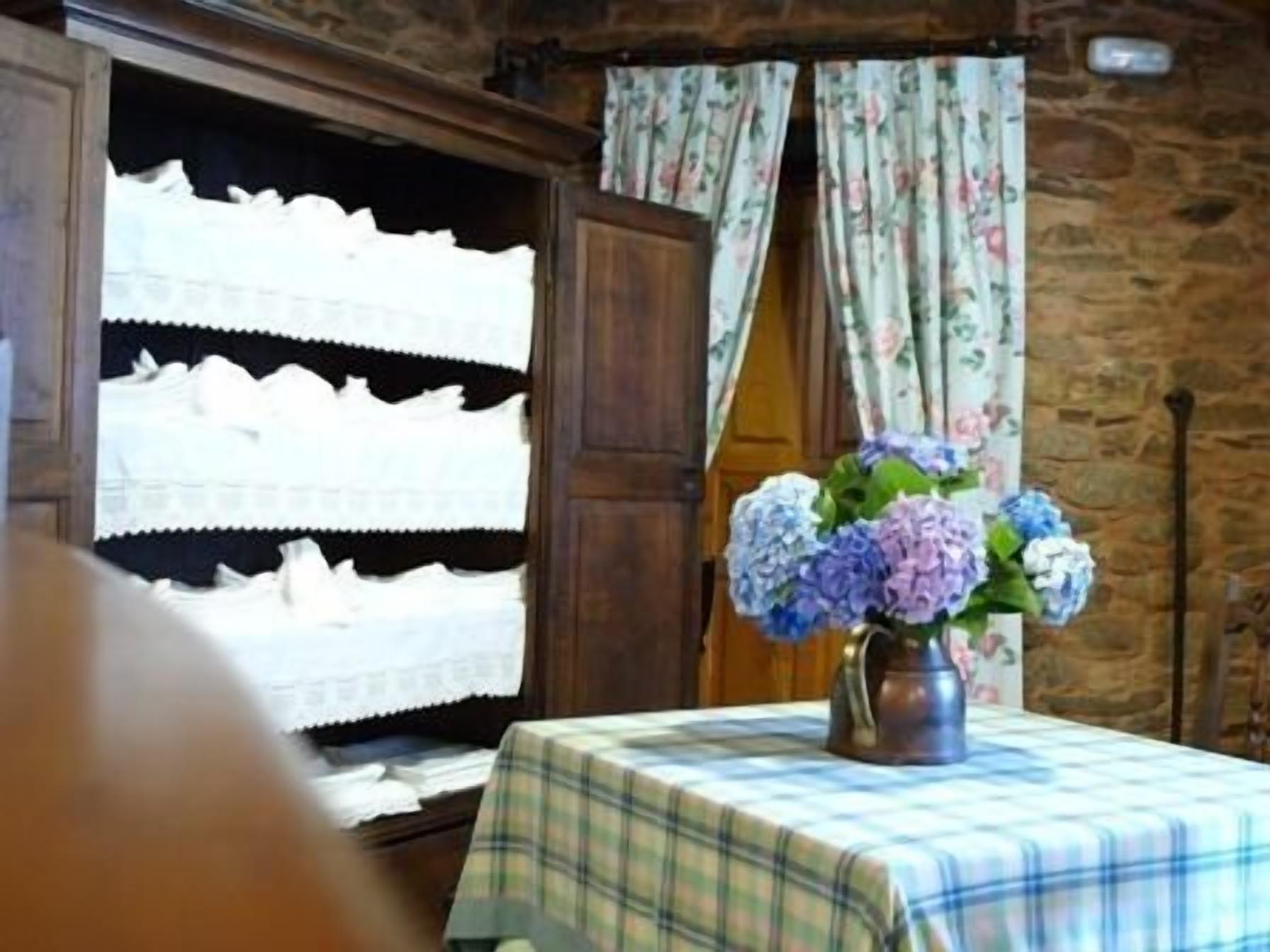 Une pièce comprenant un meuble en bois où sont exposés des articles en dentelle blanche, une table recouverte d’une nappe à carreaux et un vase rempli de fleurs.