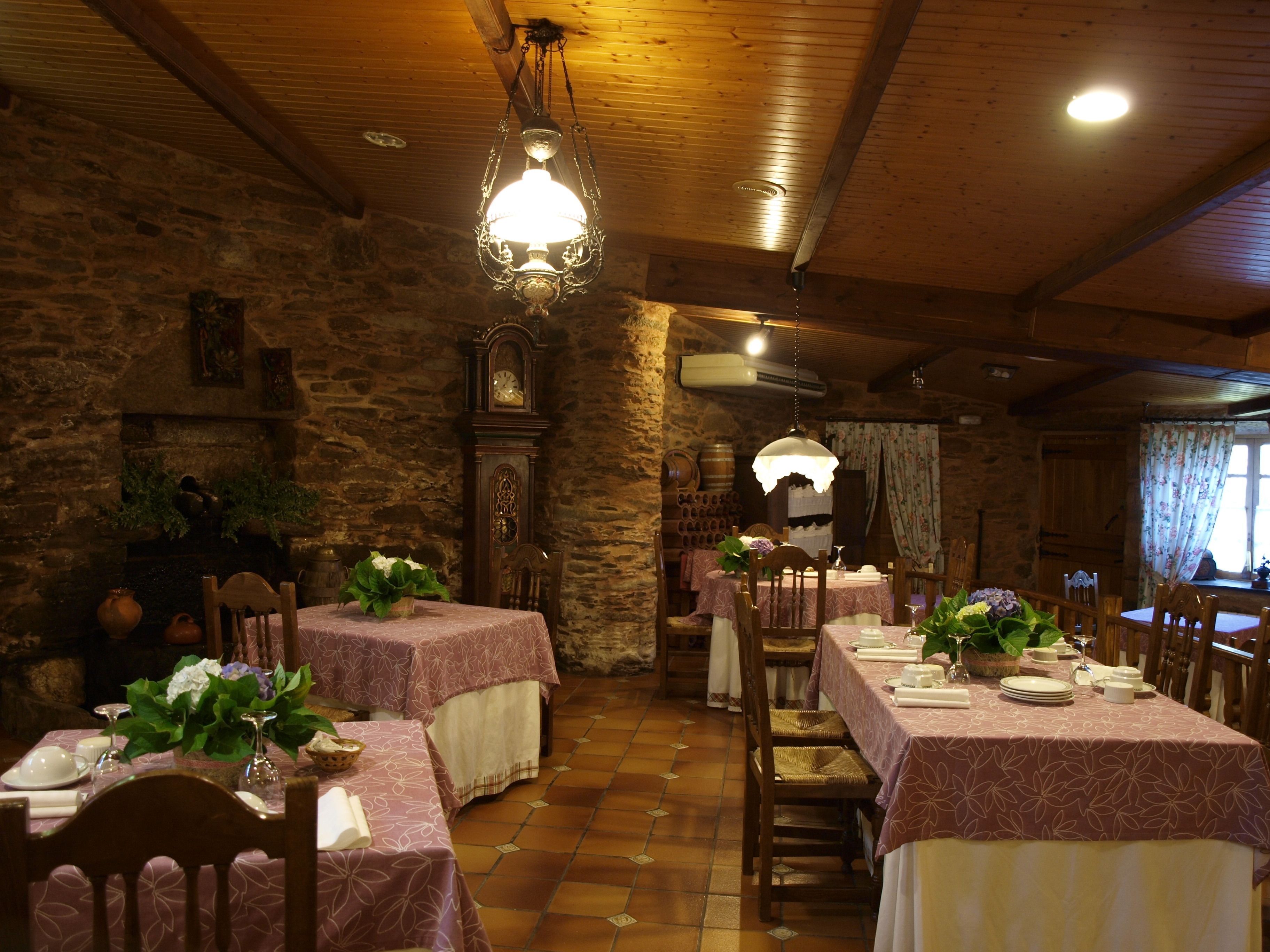 A rustic dining area with stone walls, wooden beams, and a chandelier. Tables are set with white tablecloths and floral centerpieces.