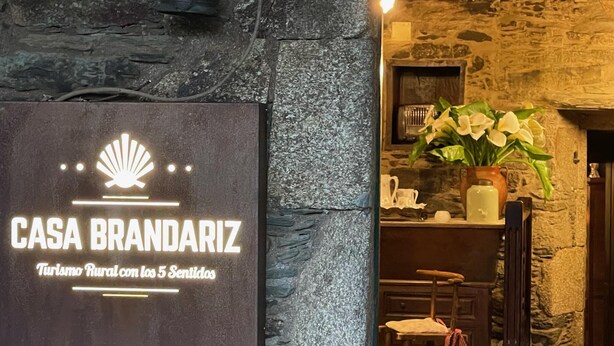 A stone wall with a sign for 'CASA BRANDARIZ' and a rustic interior with a plant and wooden furniture. - Casa Brandariz (Arzua)