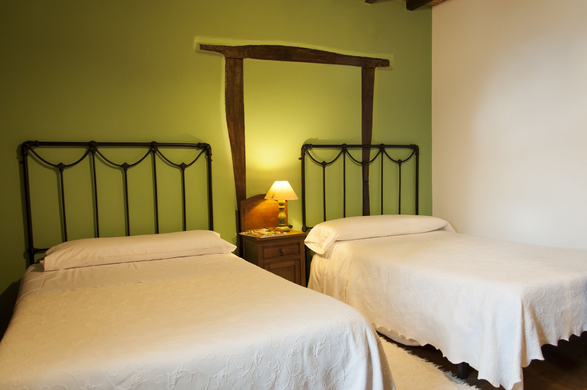 A room with two beds, each with a wooden headboard and white bedding. A nightstand with a lamp is between the beds.