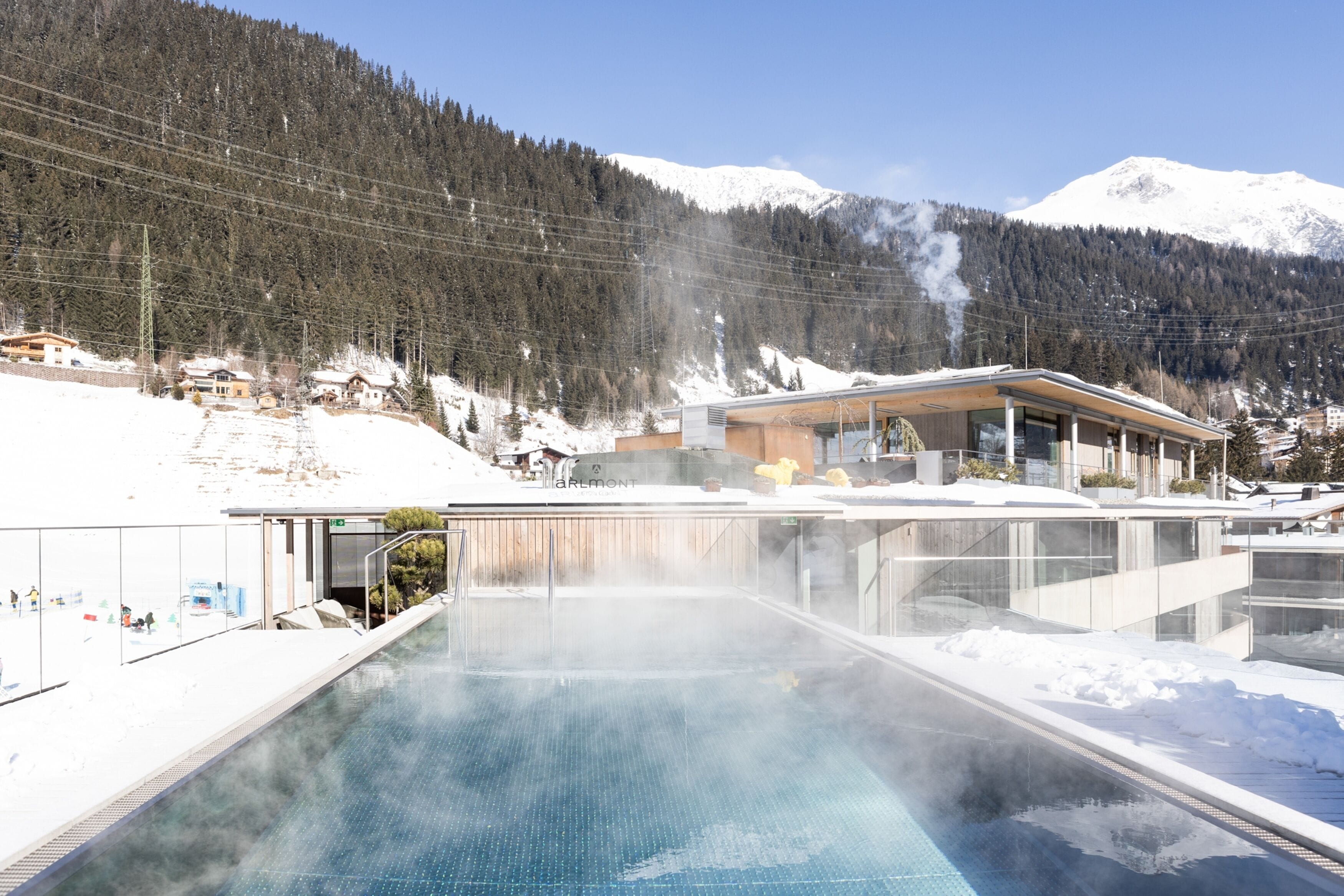 Um spa moderno com piscina envidraçada, cercado por montanhas nevadas e céu limpo.