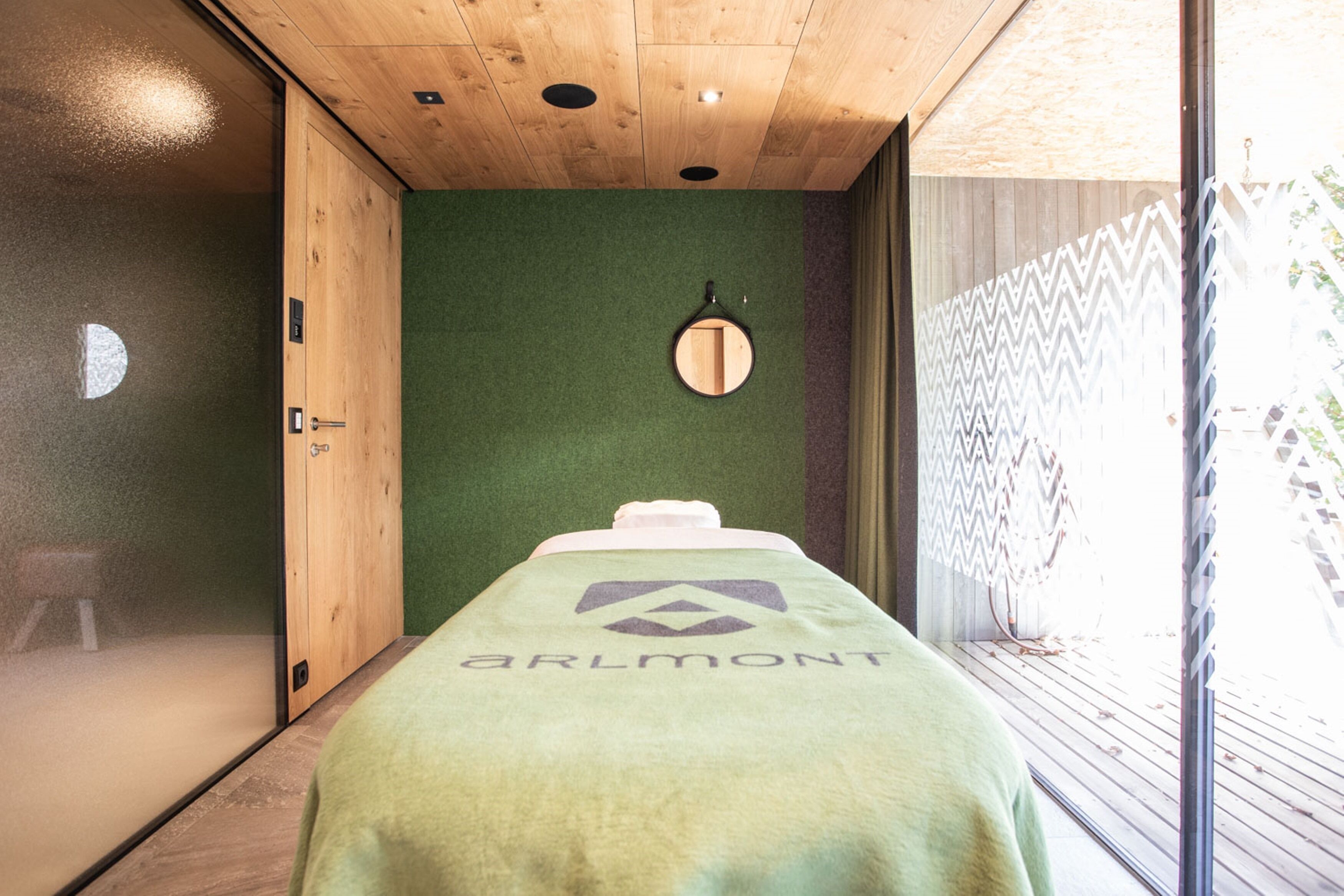 Una habitación con techo de madera, pared verde, espejo redondo y cama con manta que muestra un logotipo.