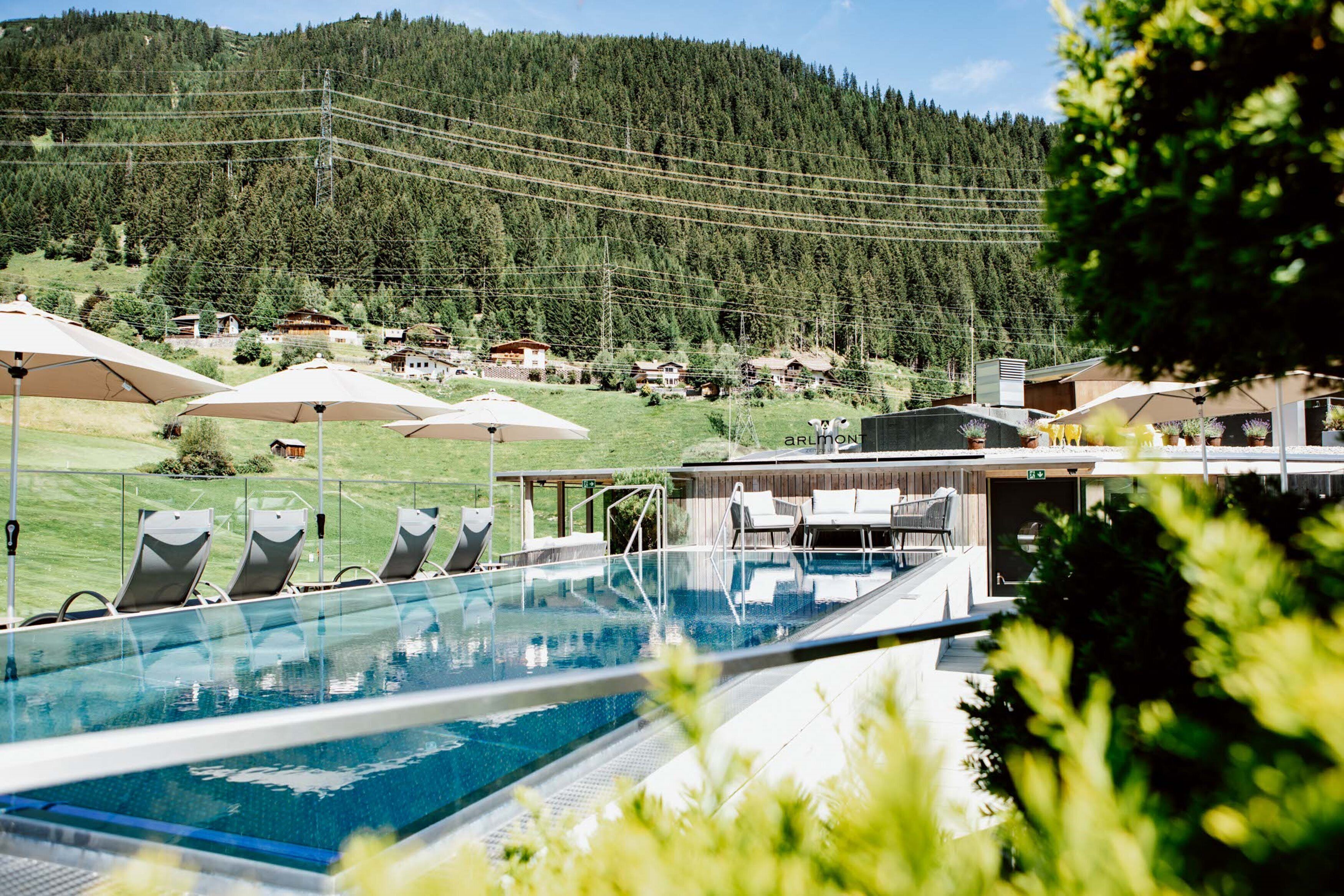 Un espace piscine doté de chaises longues et de parasols, entouré d’un paysage montagneux et de maisons.