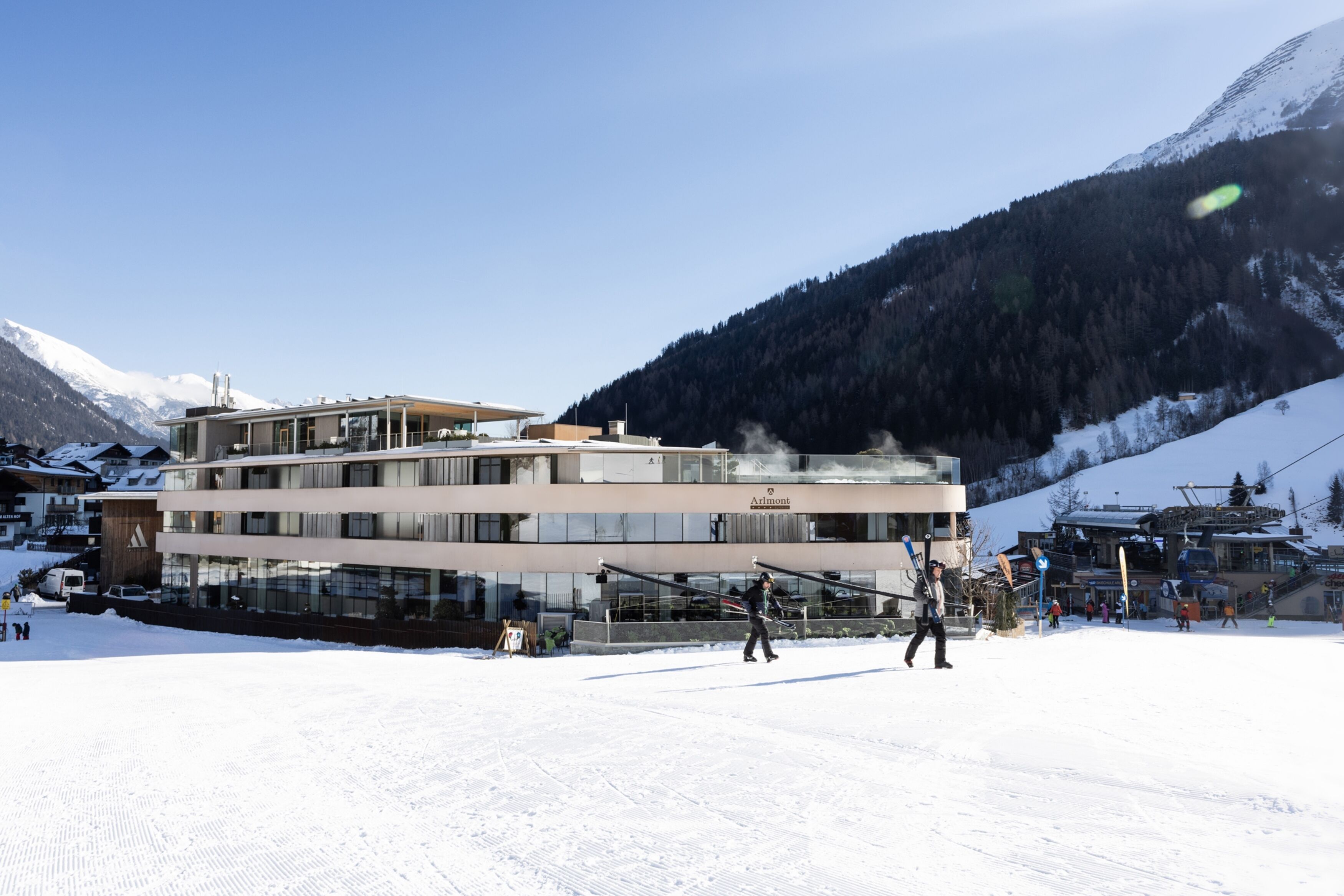 Un hôtel moderne à la façade vitrée, situé dans une région montagneuse enneigée, avec des pistes de ski visibles au loin.
