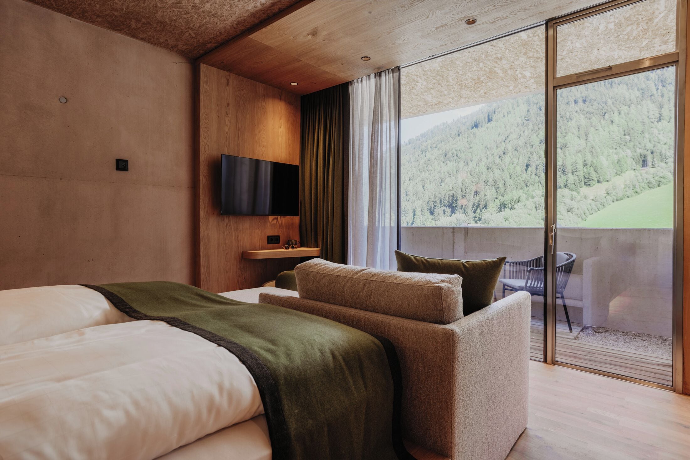 Habitación de hotel moderna con una cama grande, televisor de pantalla plana, una mesita y balcón con vistas a una zona boscosa.