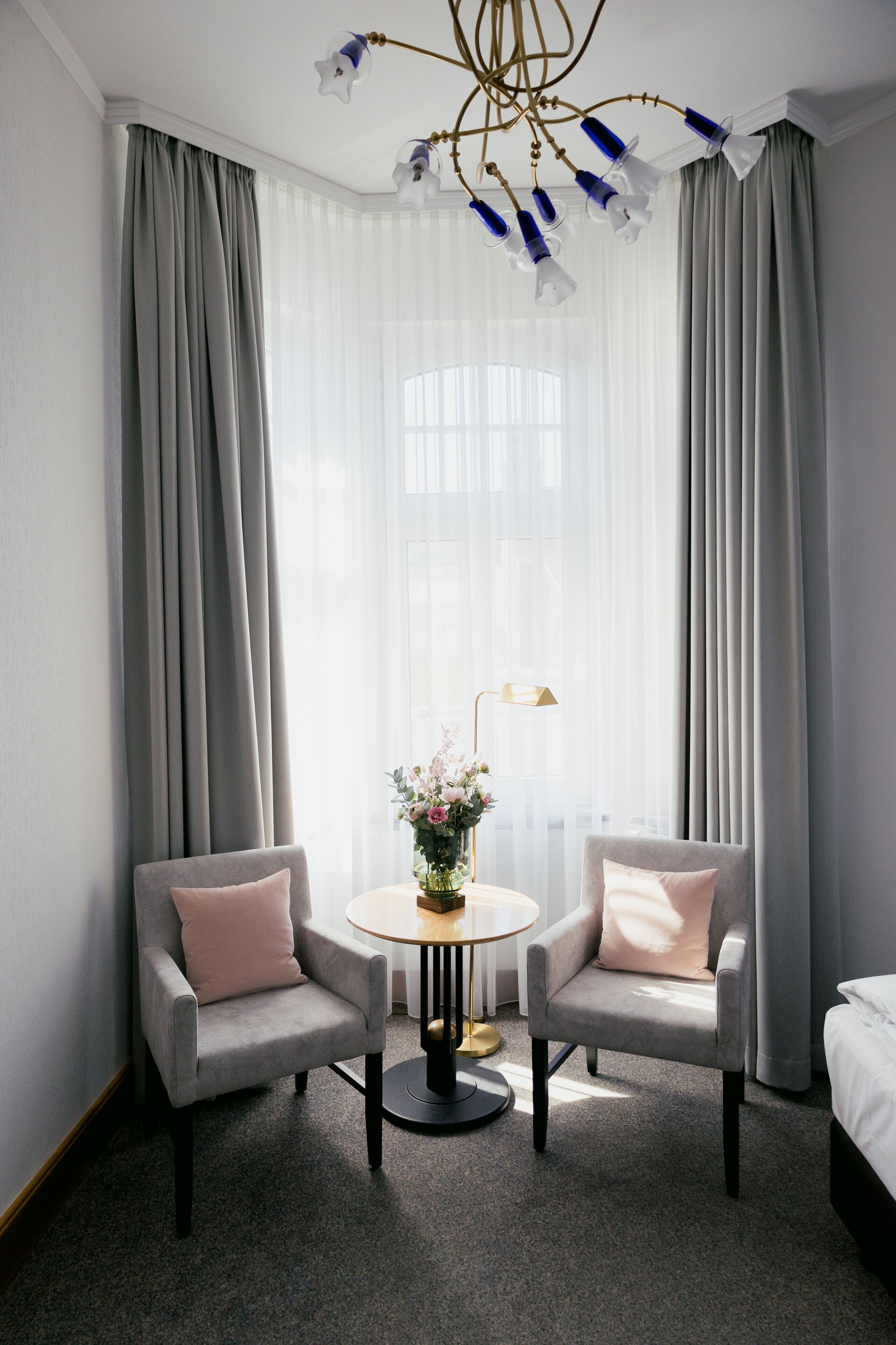 A hotel room with a large window, two armchairs, a small round table with a vase of flowers, and a bed.