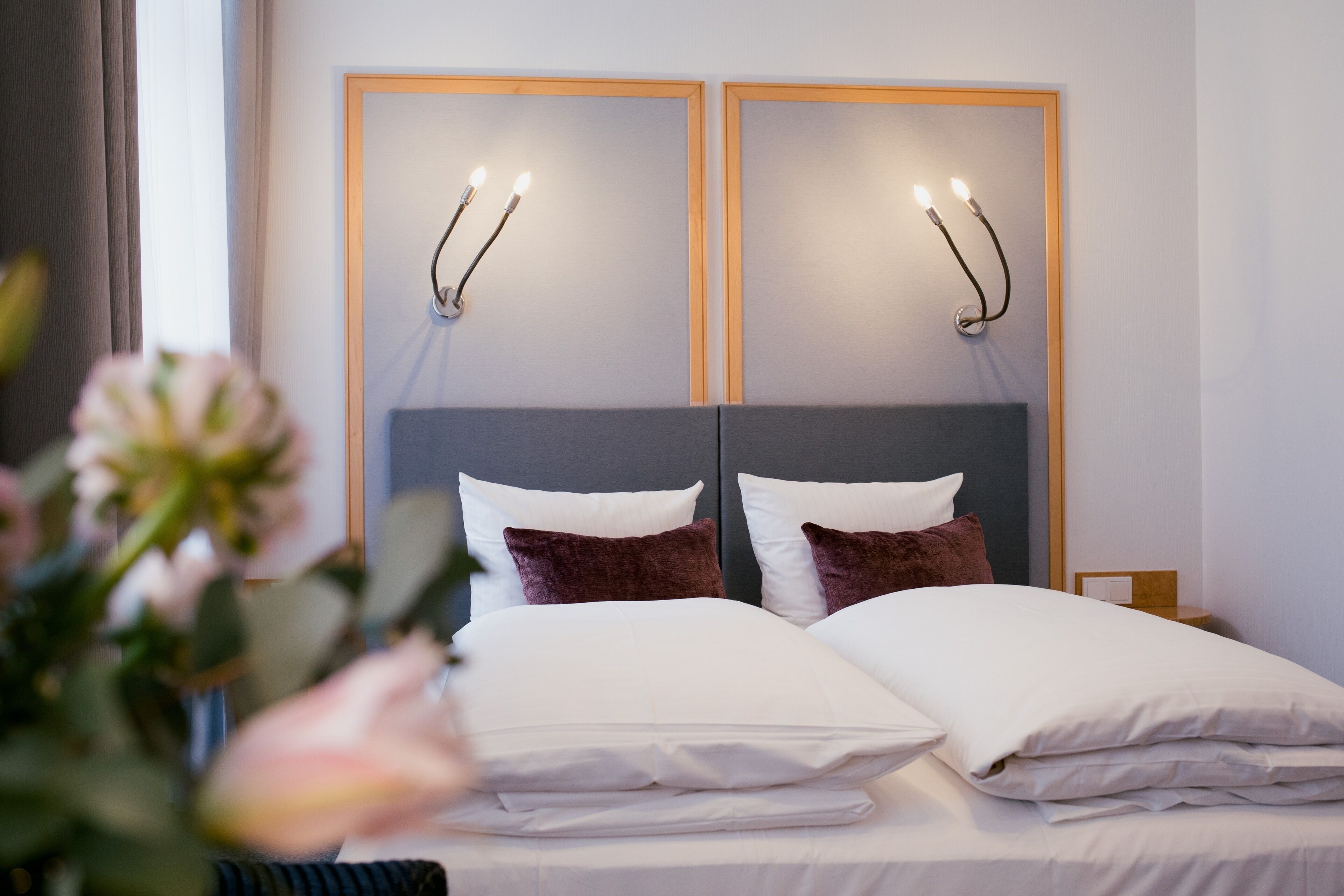 Una cama doble con almohadas blancas y marrones, un cabecero gris y dos lámparas fijadas a la pared.