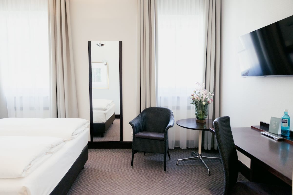 Een hotelkamer met een bed, een stoel, een kleine tafel met een bloevas, een spiegel en een aan de muur bevestigde televisie.