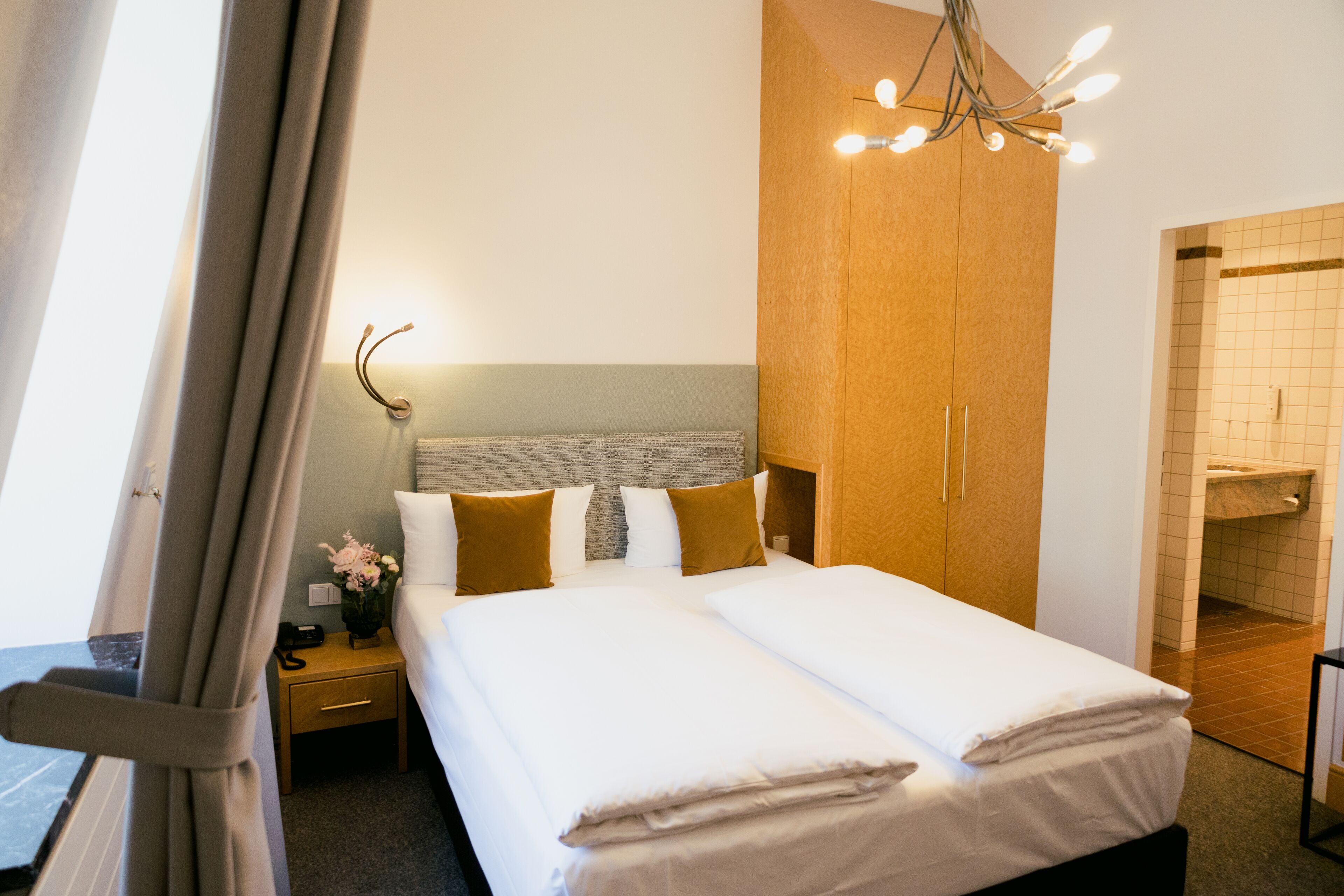 Una cama doble con sábanas blancas, cabecera de madera y mesita de noche con lámpara y un jarrón de flores.