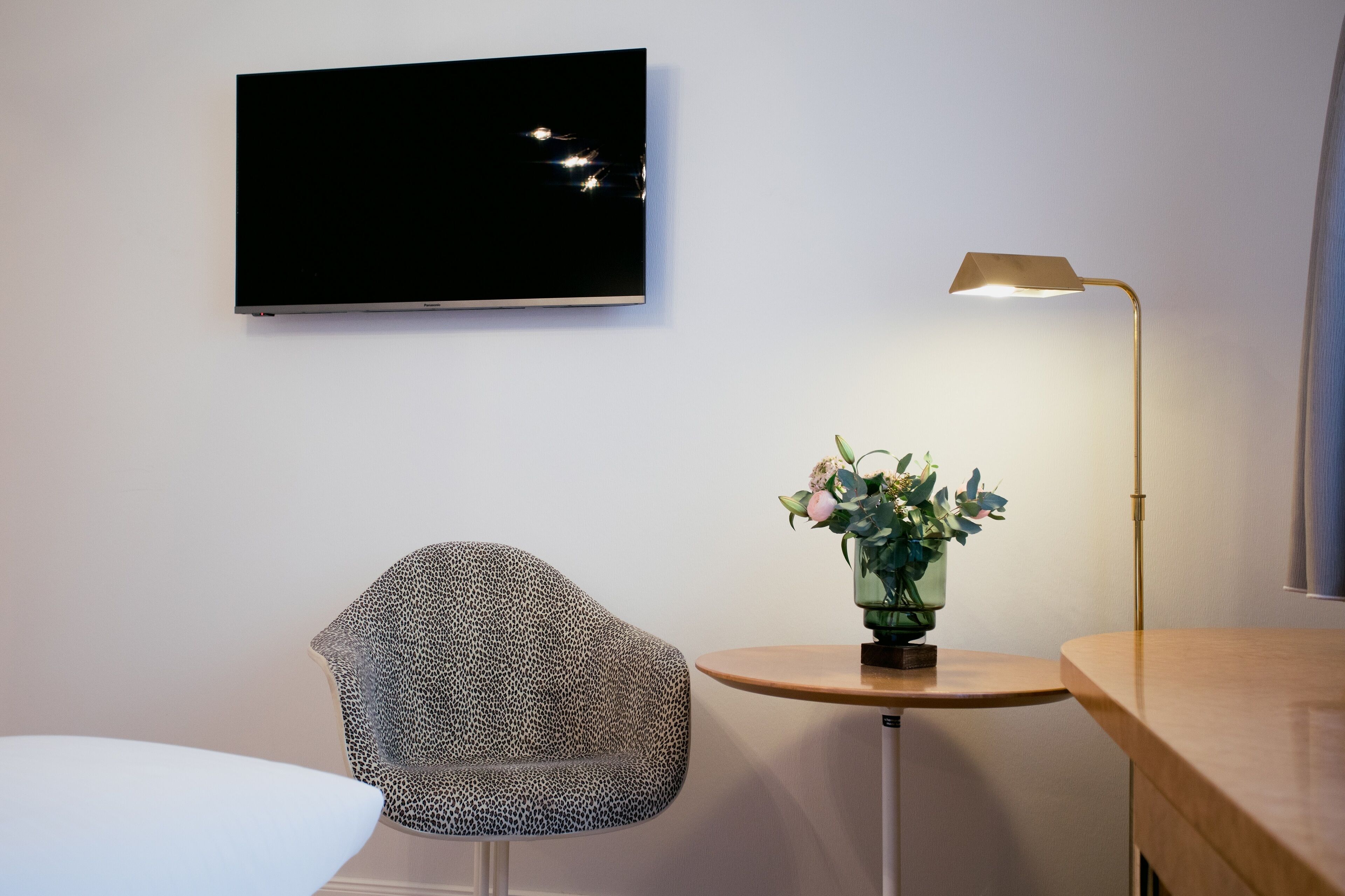 A modern room with a flat-screen TV mounted on the wall, a unique patterned chair, a small round table with a vase of flowers, and a floor lamp.