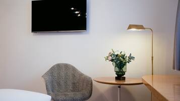 A modern room with a flat-screen TV mounted on the wall, a unique patterned chair, a small round table with a vase of flowers, and a floor lamp.