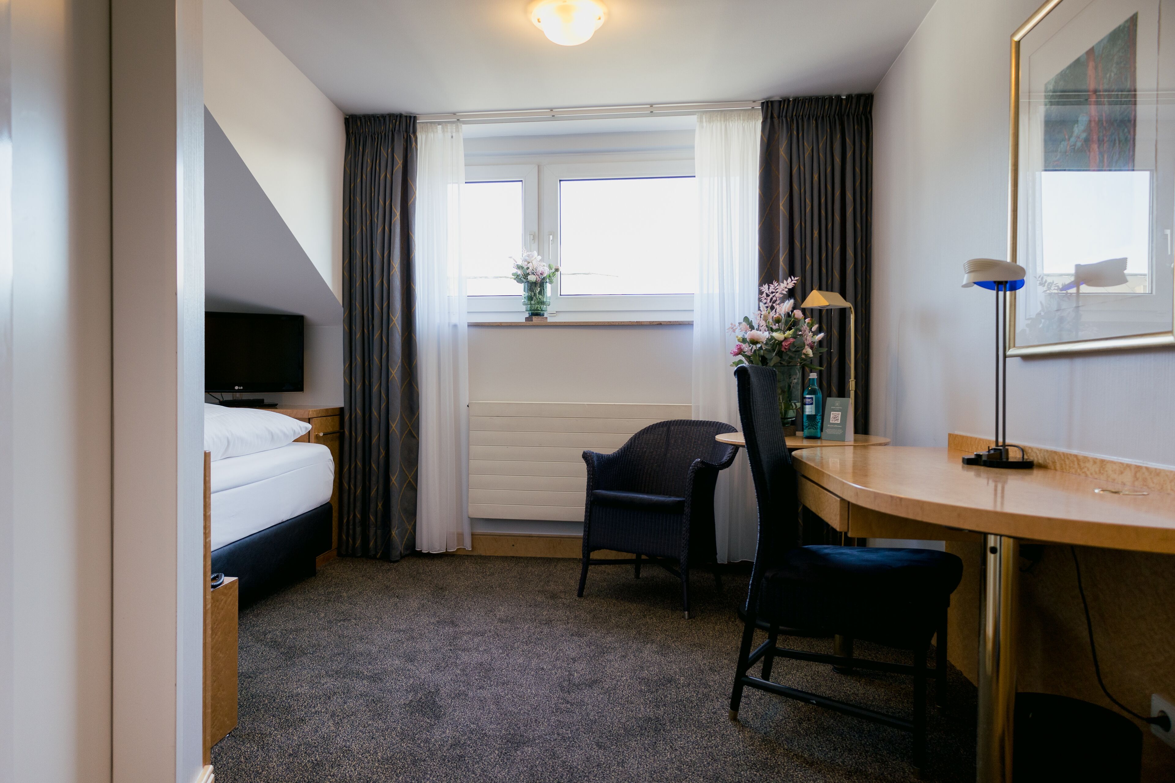 A hotel room with a bed, a desk, a chair, a TV, a window with curtains, and a vase with flowers.