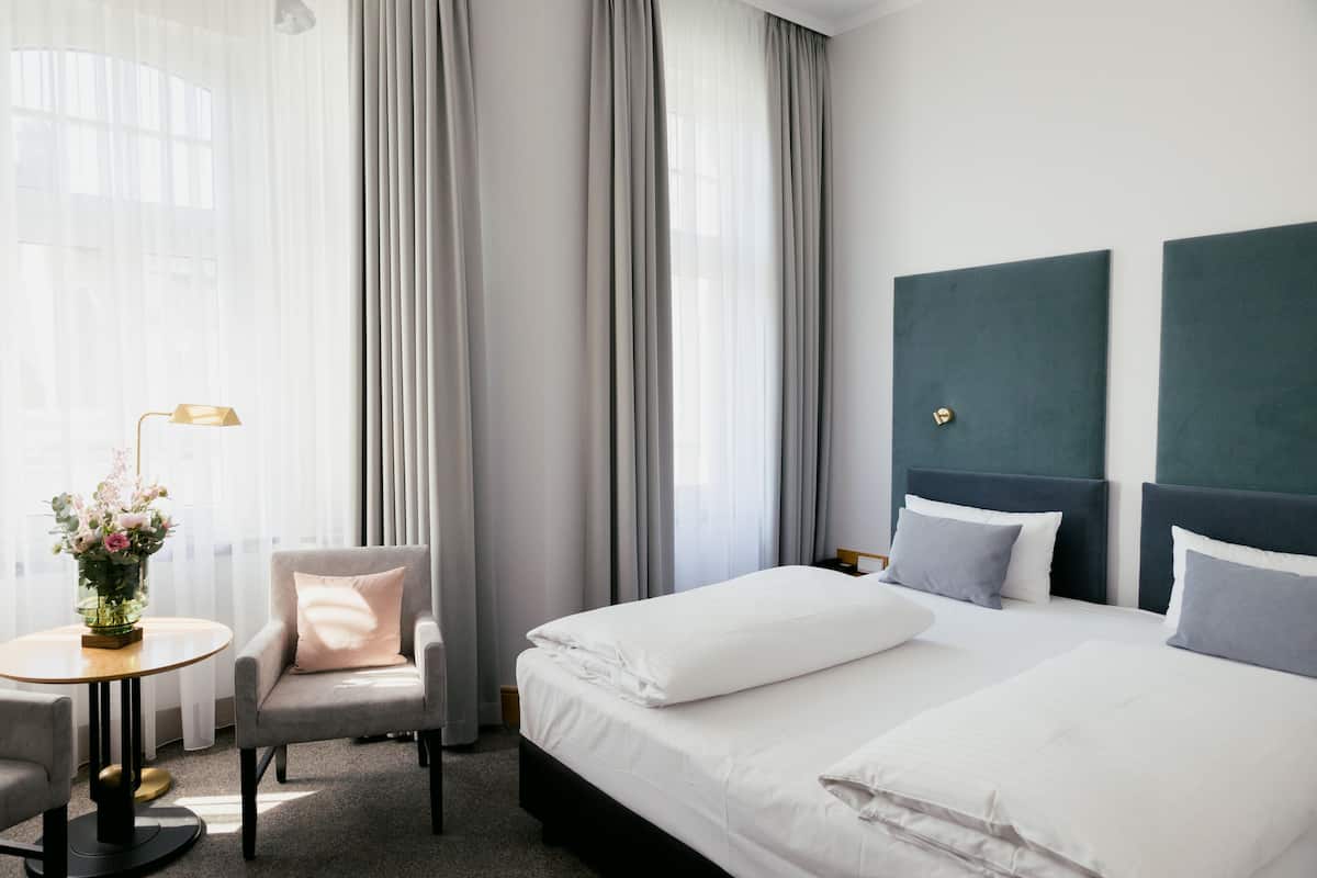 Een hotelkamer met een bed, een stoel, een klein tafeltje met een lamp en een raam met gordijnen.