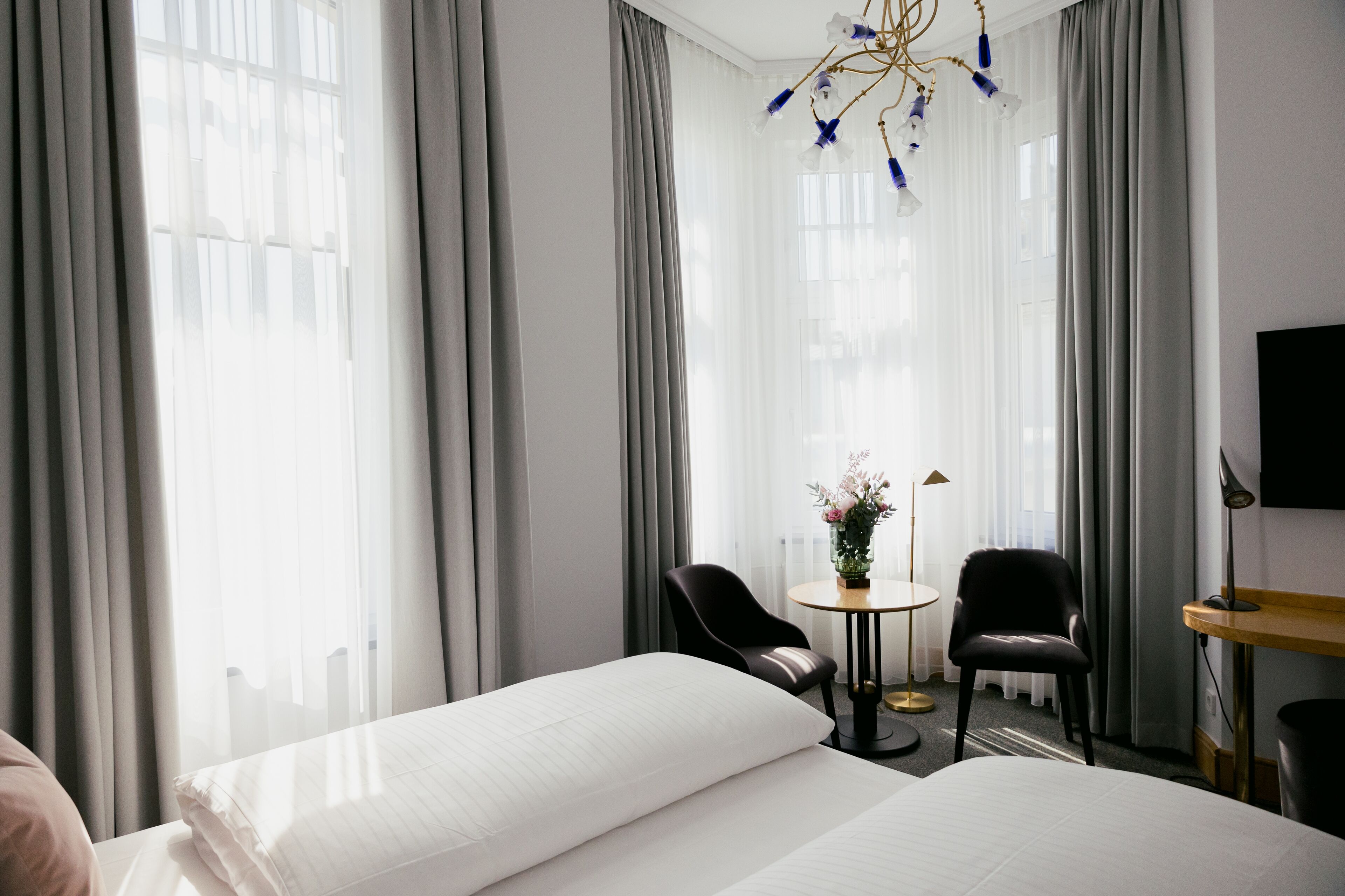 Habitación de hotel con dos camas, una mesita redonda con sillas, un candelabro y ventanas amplias con cortinas.