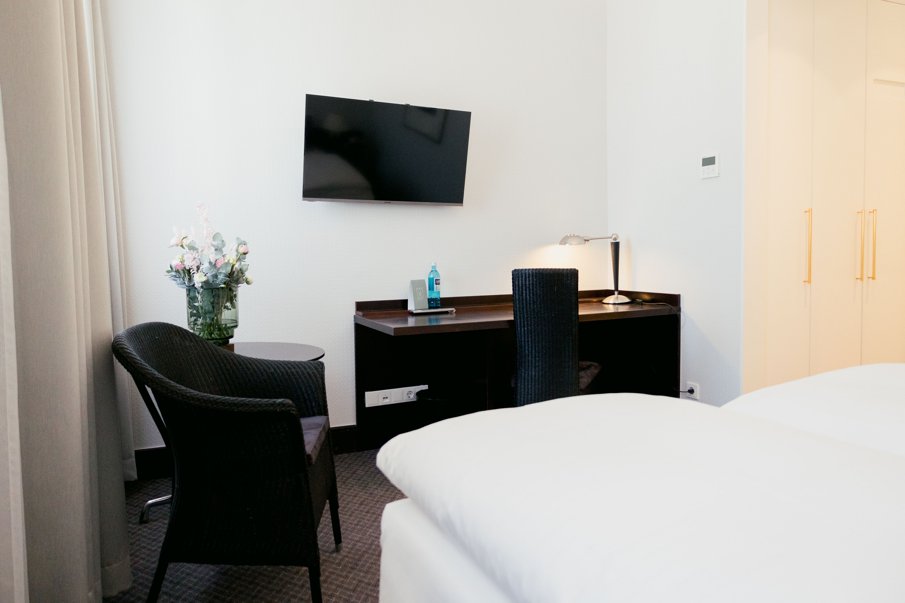 A hotel room with a bed, a desk with a chair, a TV mounted on the wall, and a lamp.