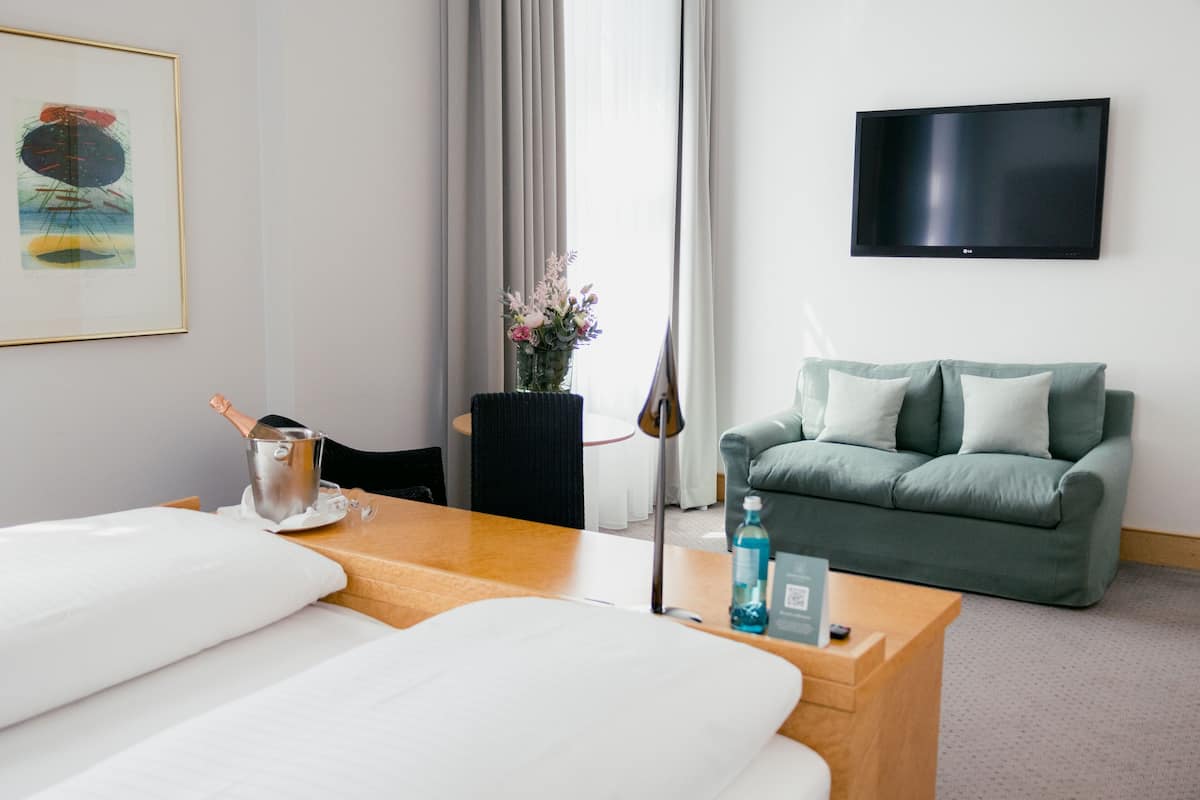 Een hotelkamer met een bed, een houten nachtkastje, een groene bank, een aan de muur bevestigde televisie en een foto in een lijst.