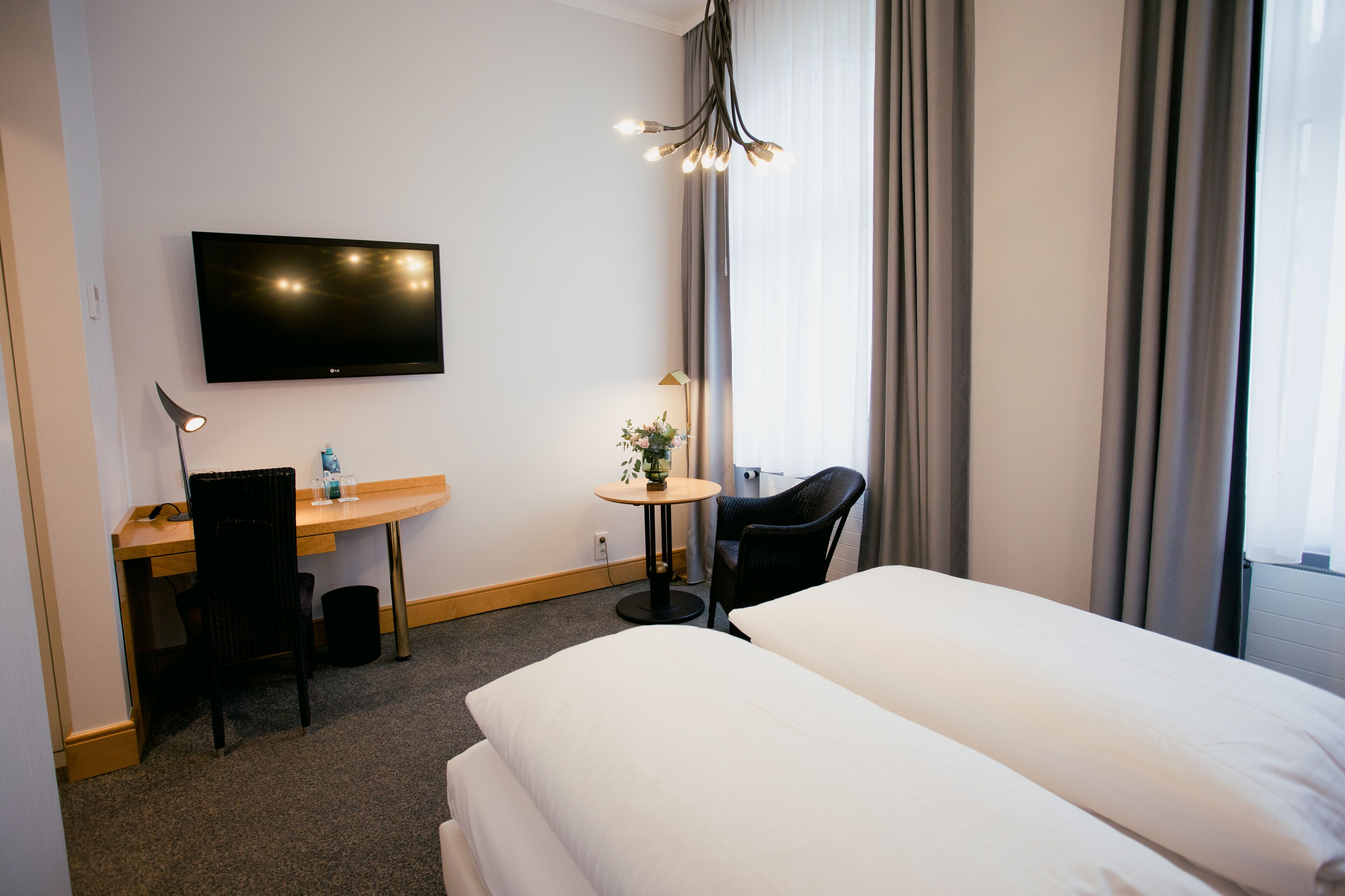 Ein Hotelzimmer mit einem Bett, einem Schreibtisch mit Stuhl, einem Fernseher, einem kleinen Tisch mit Pflanze und einem Fenster mit Vorhängen.