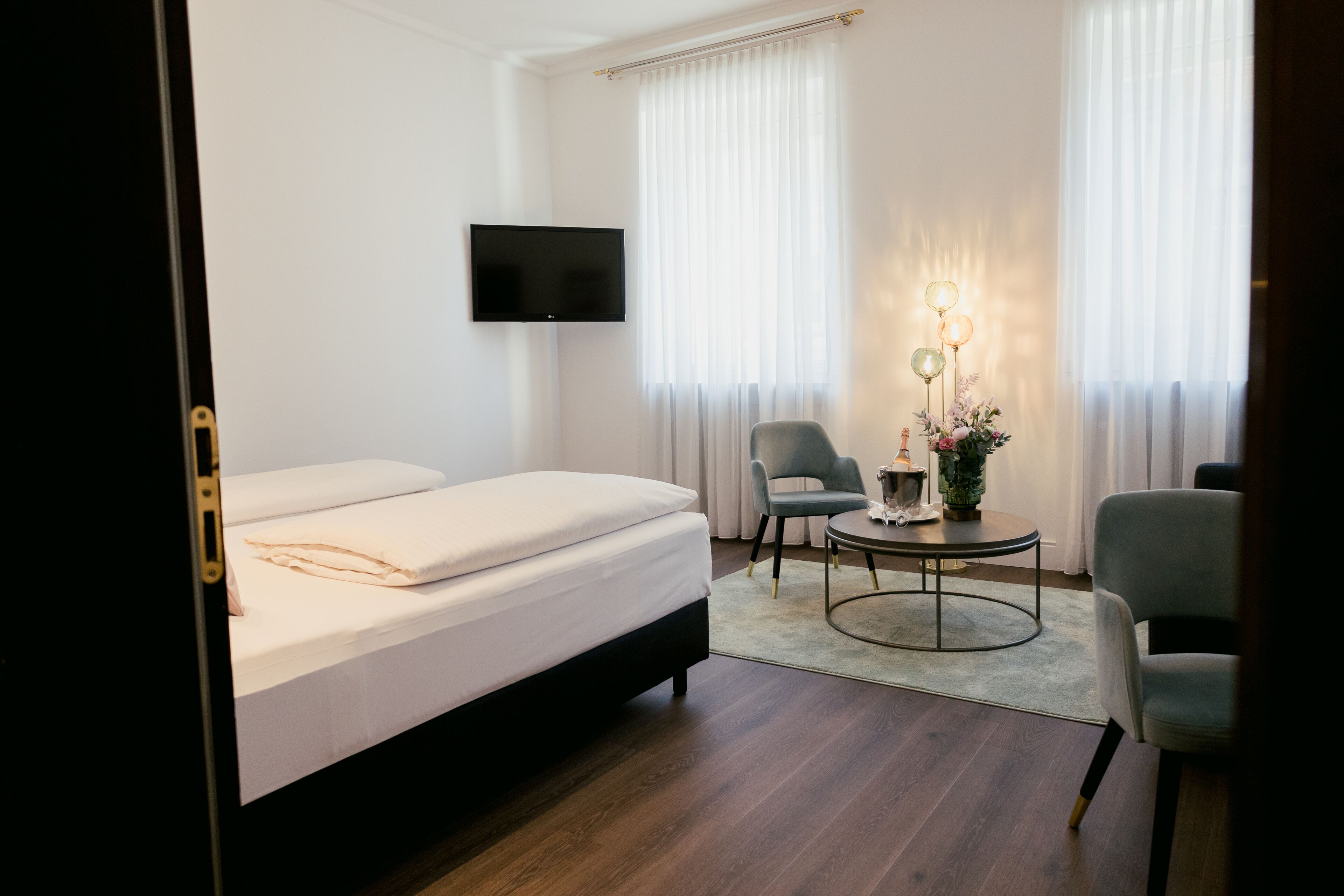 A hotel room with a bed, two chairs, a small table with a lamp, and a TV mounted on the wall.