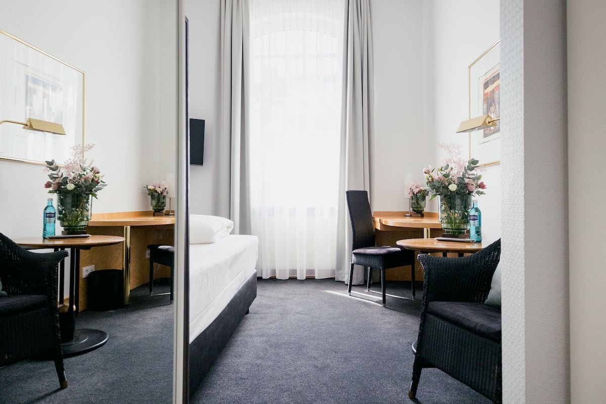 Een hotelkamer met een bed, twee stoelen, een tafel en een bureau. Op de tafel staan twee bloemstukken en er is een raam met gordijnen.