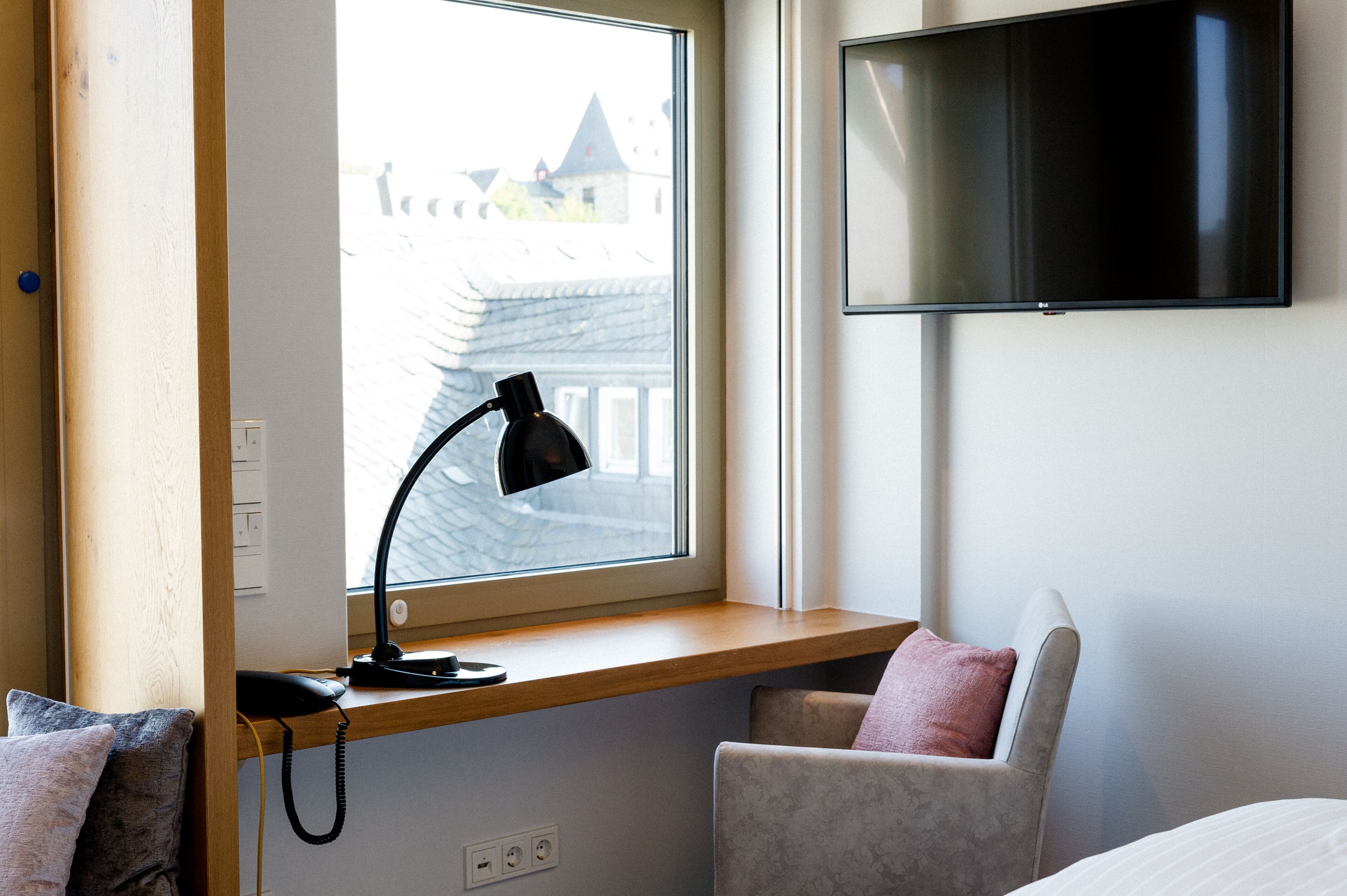 A hotel room with a desk by the window, a flat-screen TV mounted on the wall, and a chair with a cushion.