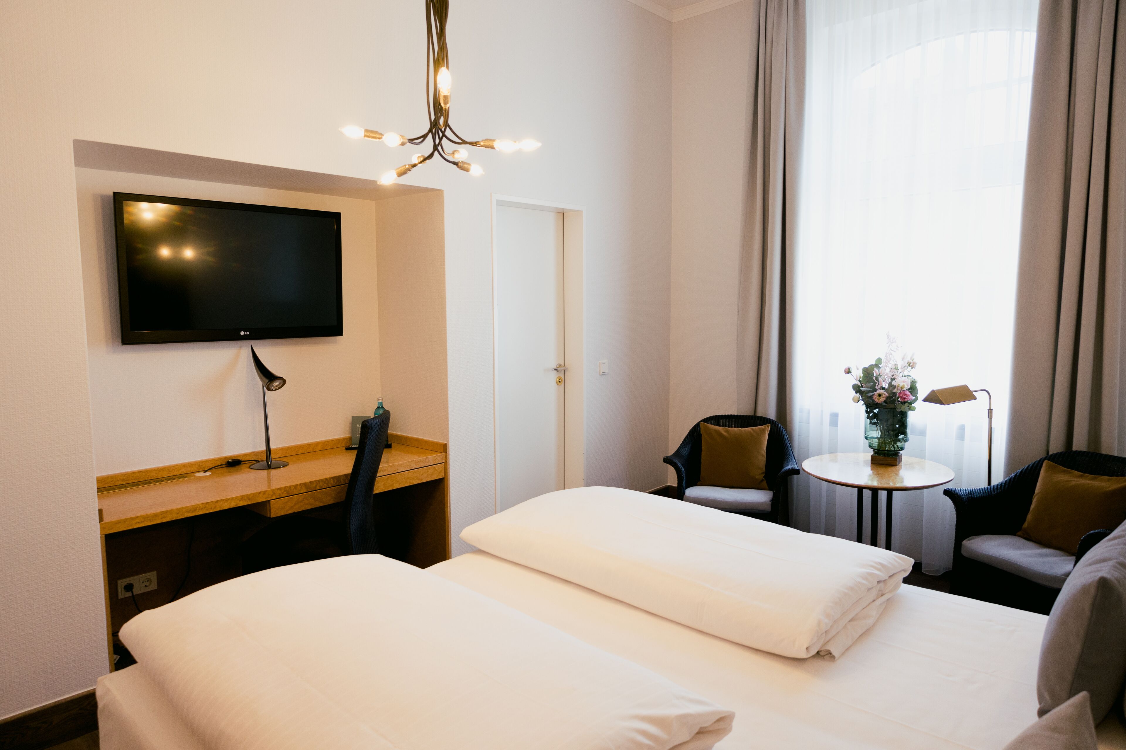 Une chambre d’hôtel équipée d’un téléviseur à écran plat fixé au mur, d’un bureau avec une chaise, de deux lits avec du linge de lit blanc, d’une petite table ronde agrémentée d’un vase de fleurs et d’un canapé.