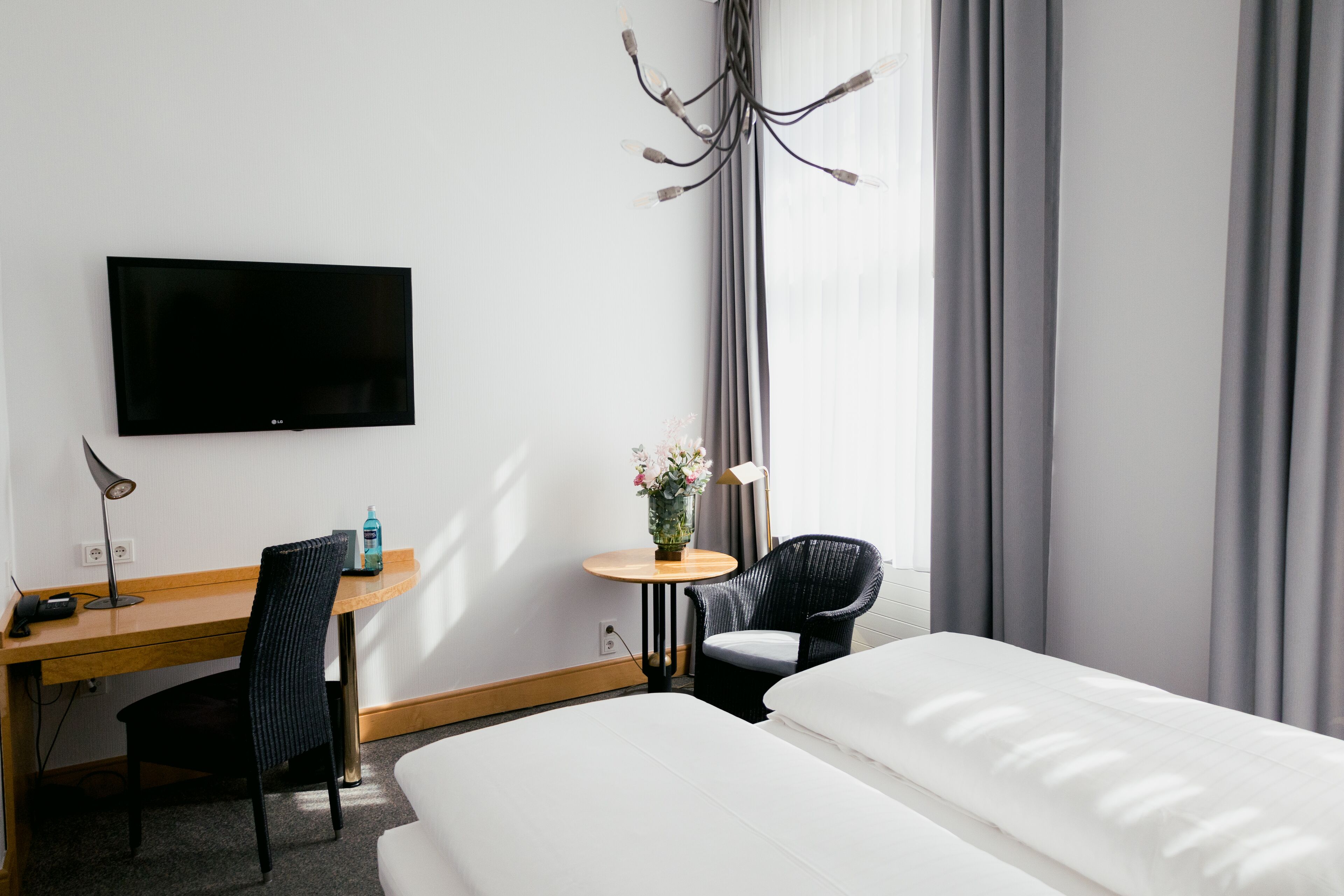 Habitación de hotel con una cama, un escritorio con una silla, un televisor, una mesita con un jarrón de flores y una ventana con cortinas.