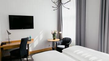 A hotel room with a bed, a desk with a chair, a television, a small table with a vase of flowers, and a window with curtains.