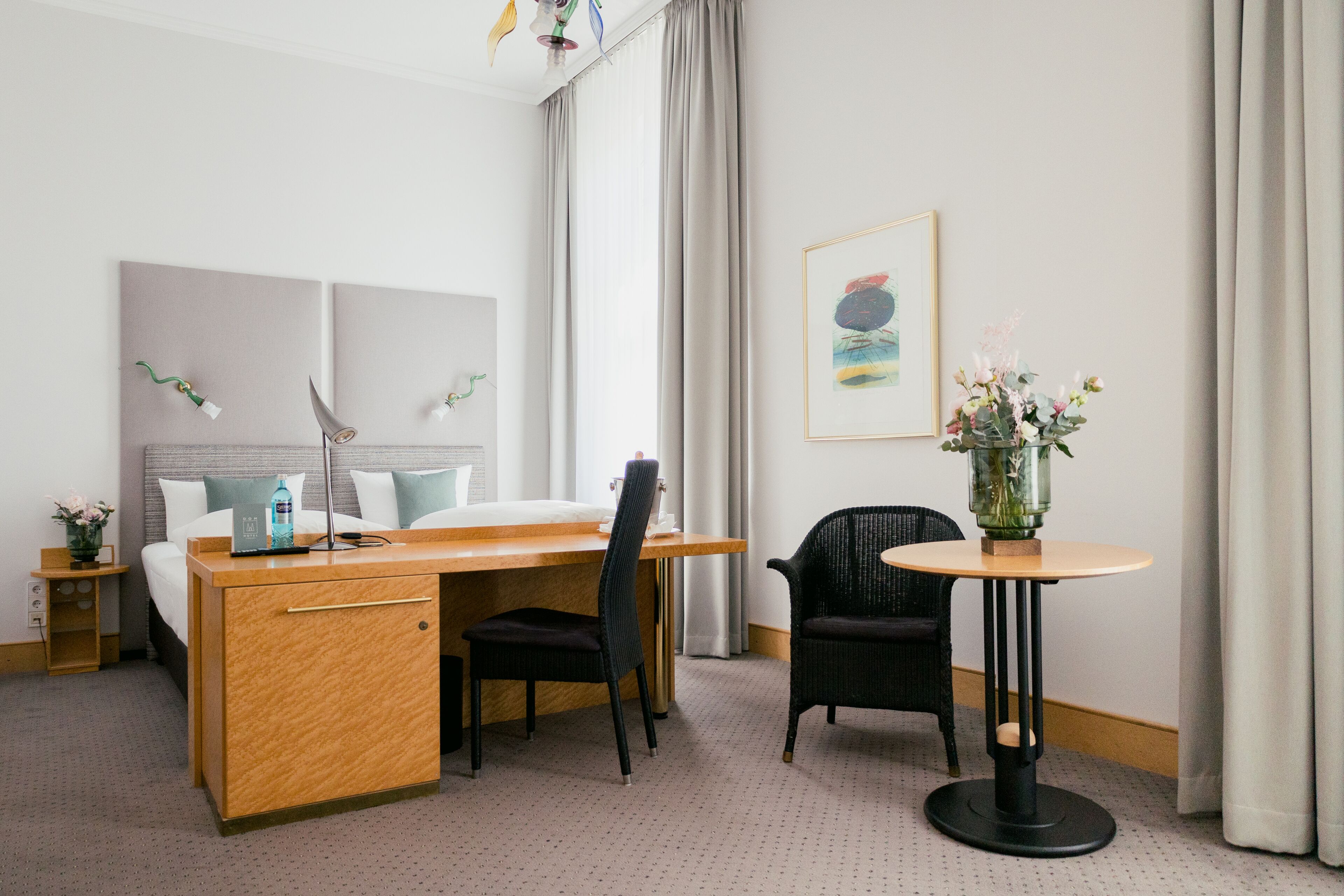 Ein Hotelzimmer mit einem Holztisch, einem Bett mit weißer Bettwäsche, einem schwarzen Stuhl, einem runden Tisch mit Blumen in einer Vase und einem gerahmten Bild an der Wand.