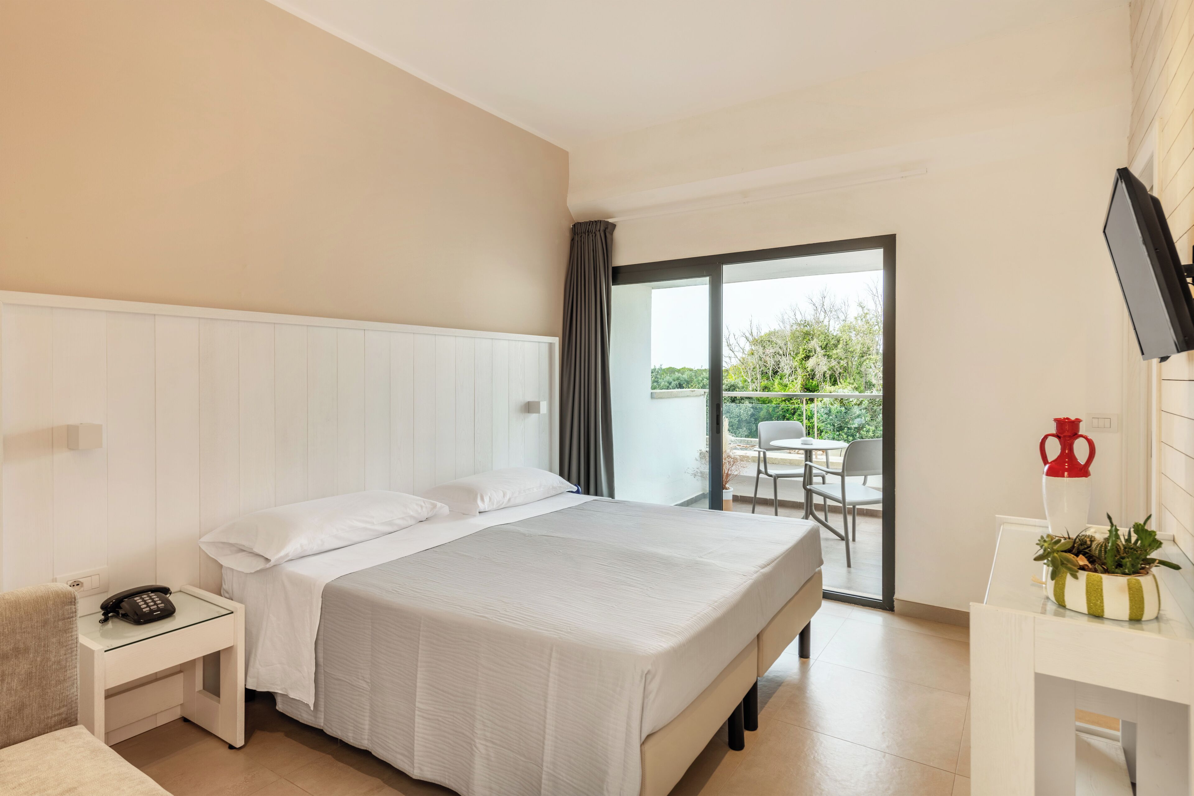 Una camera d'albergo con un letto, un comodino con il telefono, una scrivania con un vaso e piante, e una porta scorrevole in vetro che dà su uno spazio esterno.